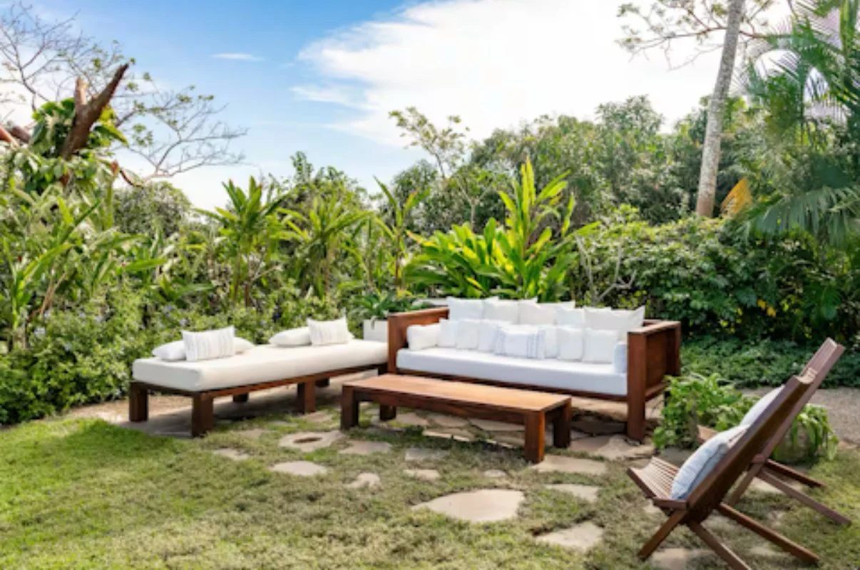 Outdoor seating area in a lush garden with wooden furniture, white cushions, and a stone pathway.