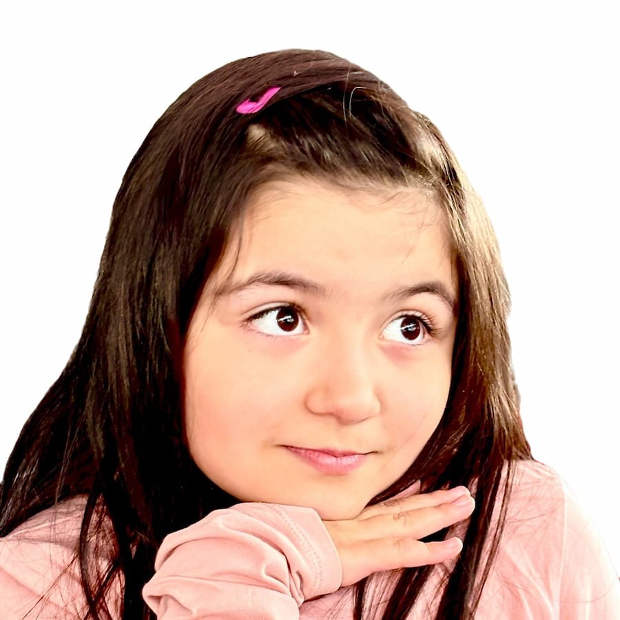 A young girl with long brown hair and a pink hair clip, wearing a pink shirt, with her chin resting on her hand, looking upward pensively.