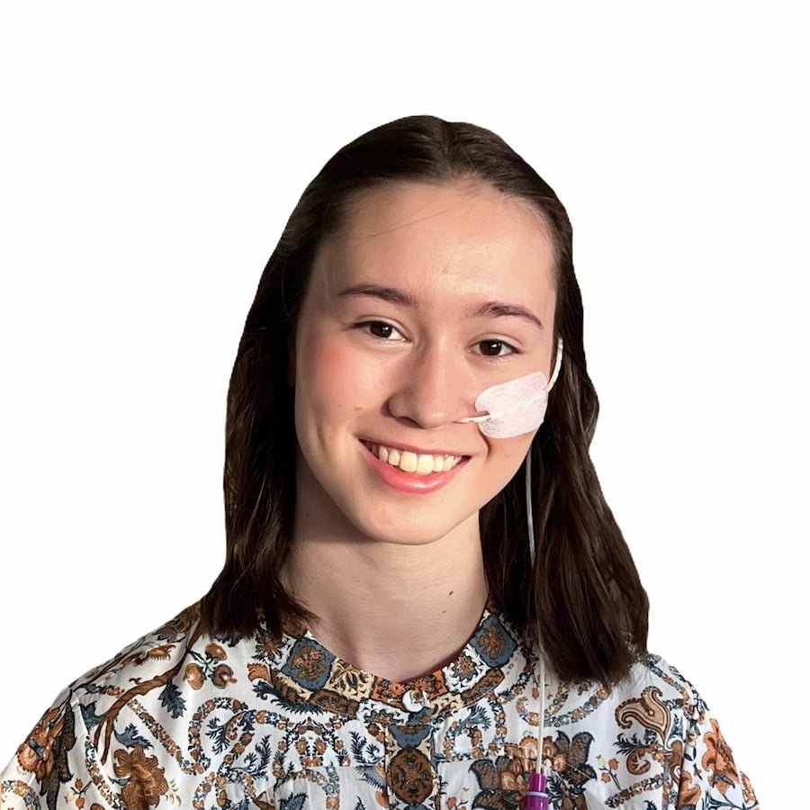 Young woman with dark hair and a ng feeding tube, wearing a patterned blouse, smiling against a white background.