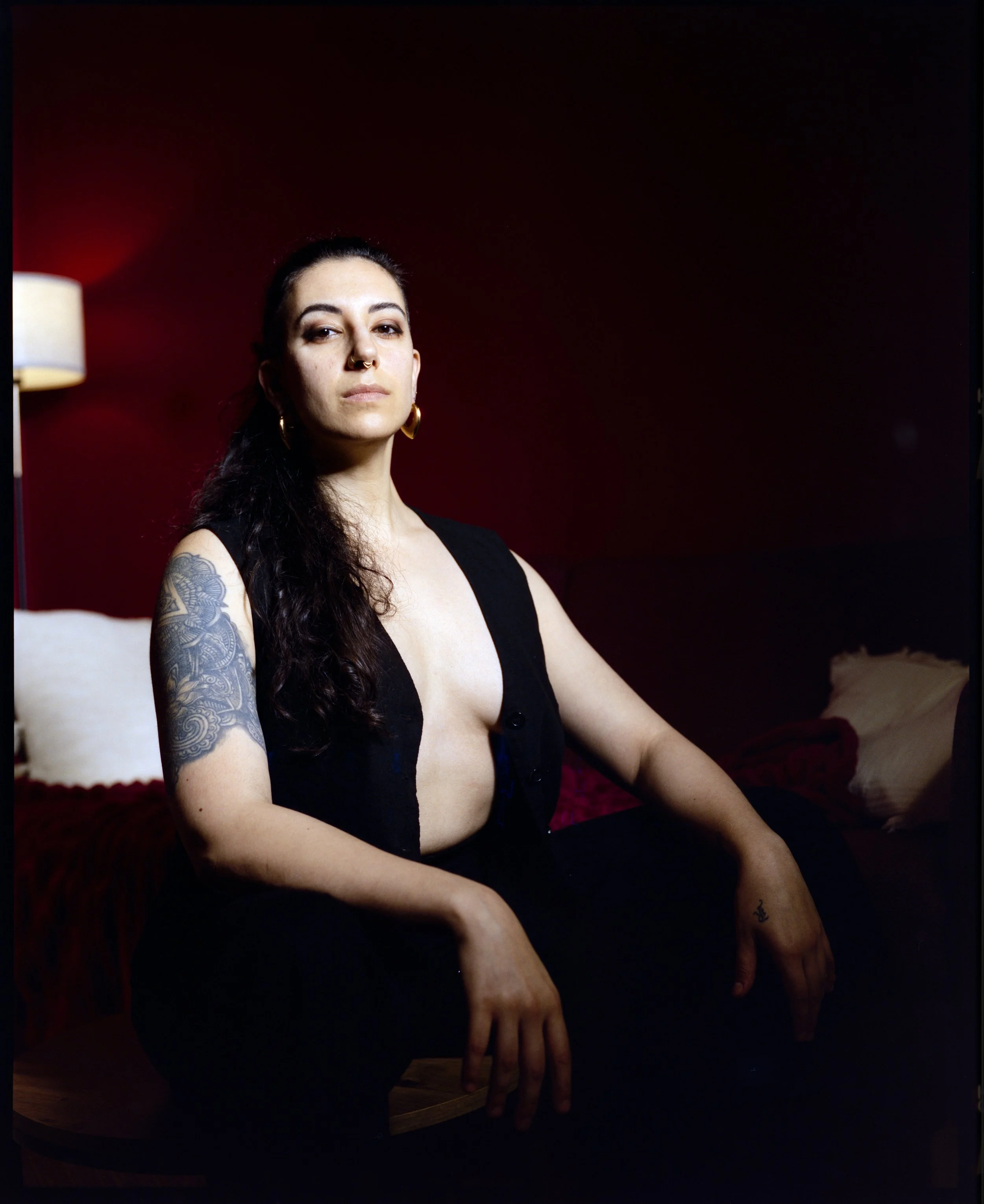 A picture of a non-binary person sitting with their arms resting on their knees. They are wearing a black open vest, black trousers and gold earrings. A tattoo covers their right upper arm.