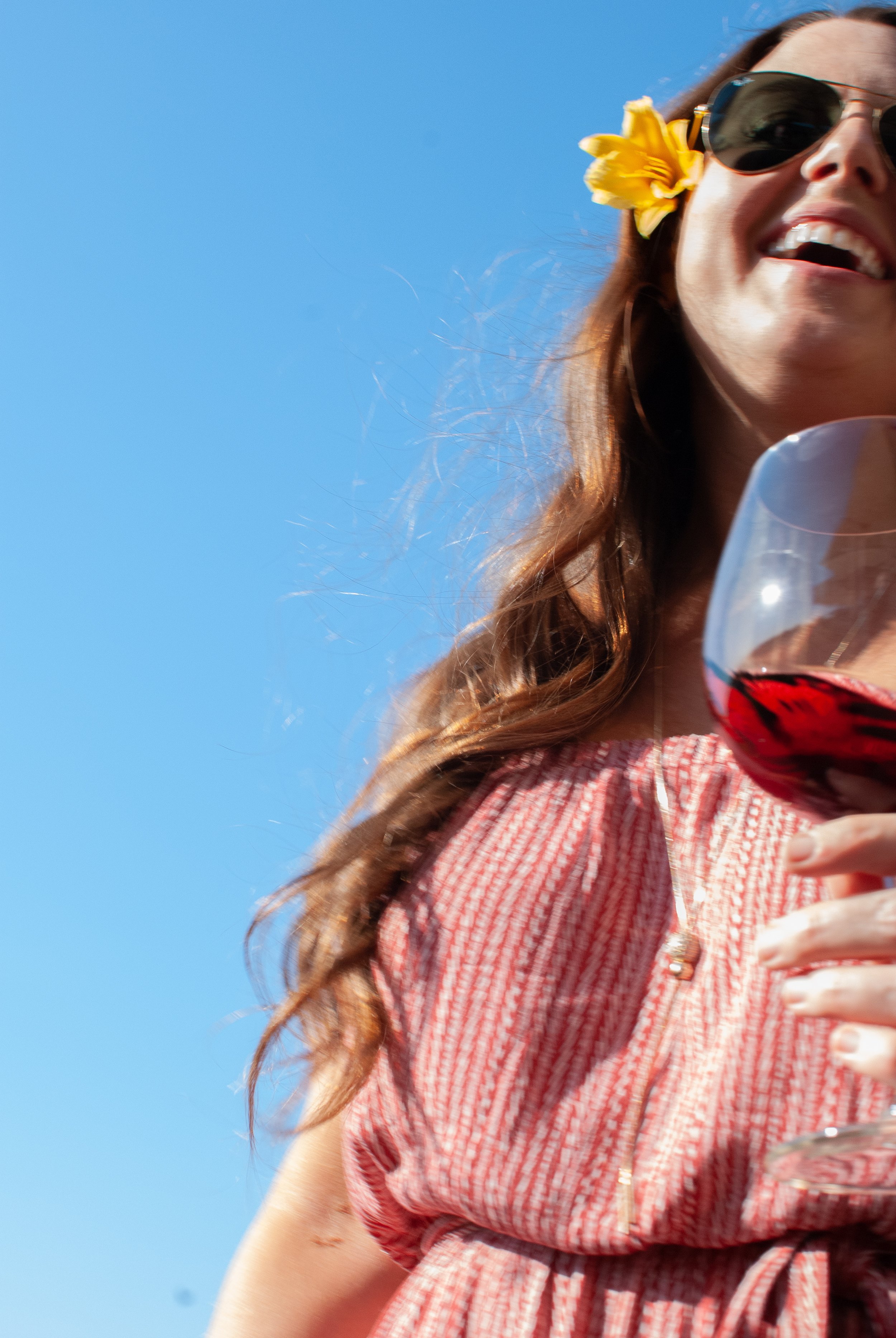 Woman smiling with sunglasses, yellow flower in hair, holding a glass of red wine, wearing a red dress against a clear blue sky.