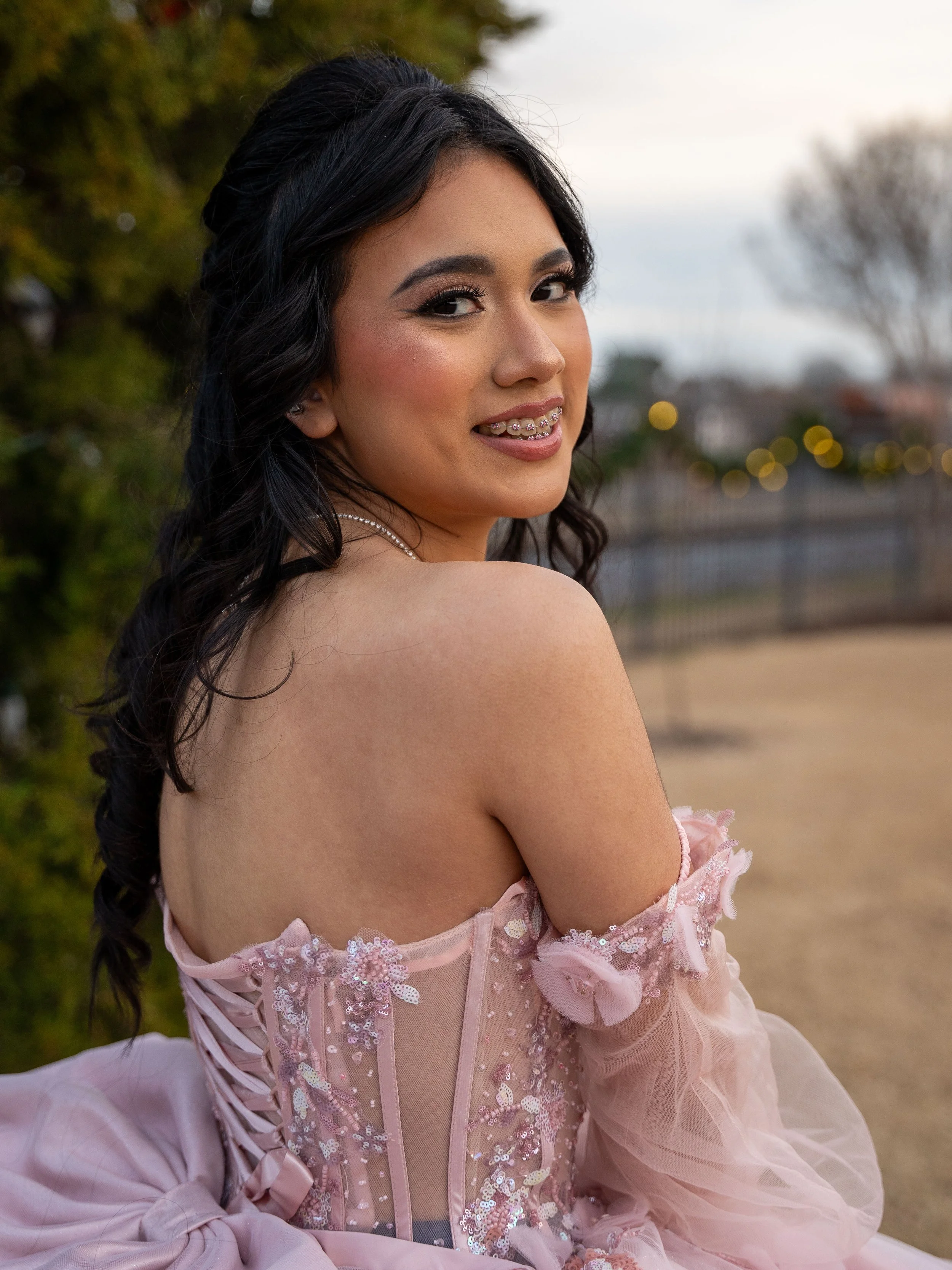 Young woman in a pink formal dress with floral embellishments and lace-up back, standing outdoors with a soft smile.