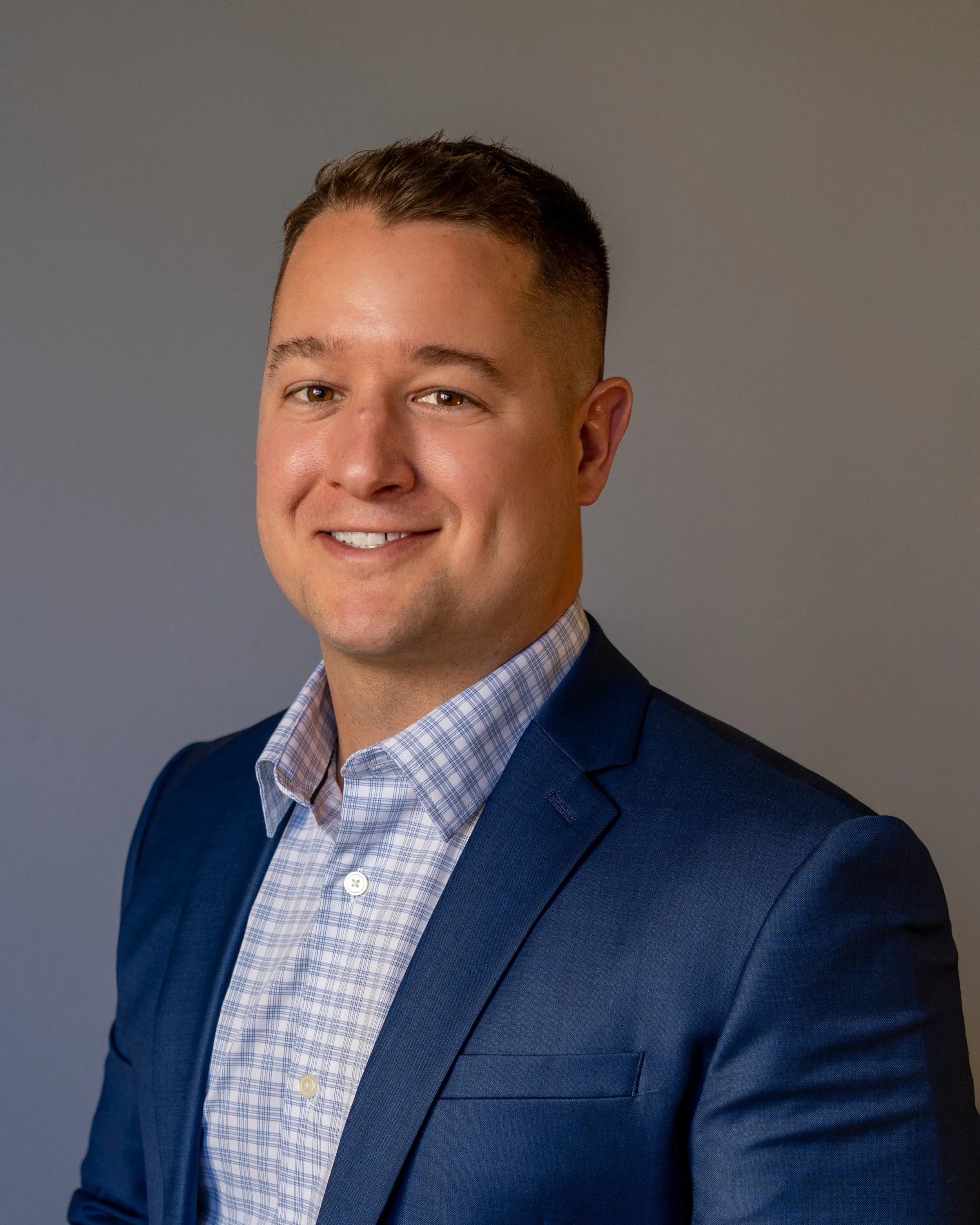Smiling man in a navy blue blazer and checkered shirt, portrait against a neutral background.