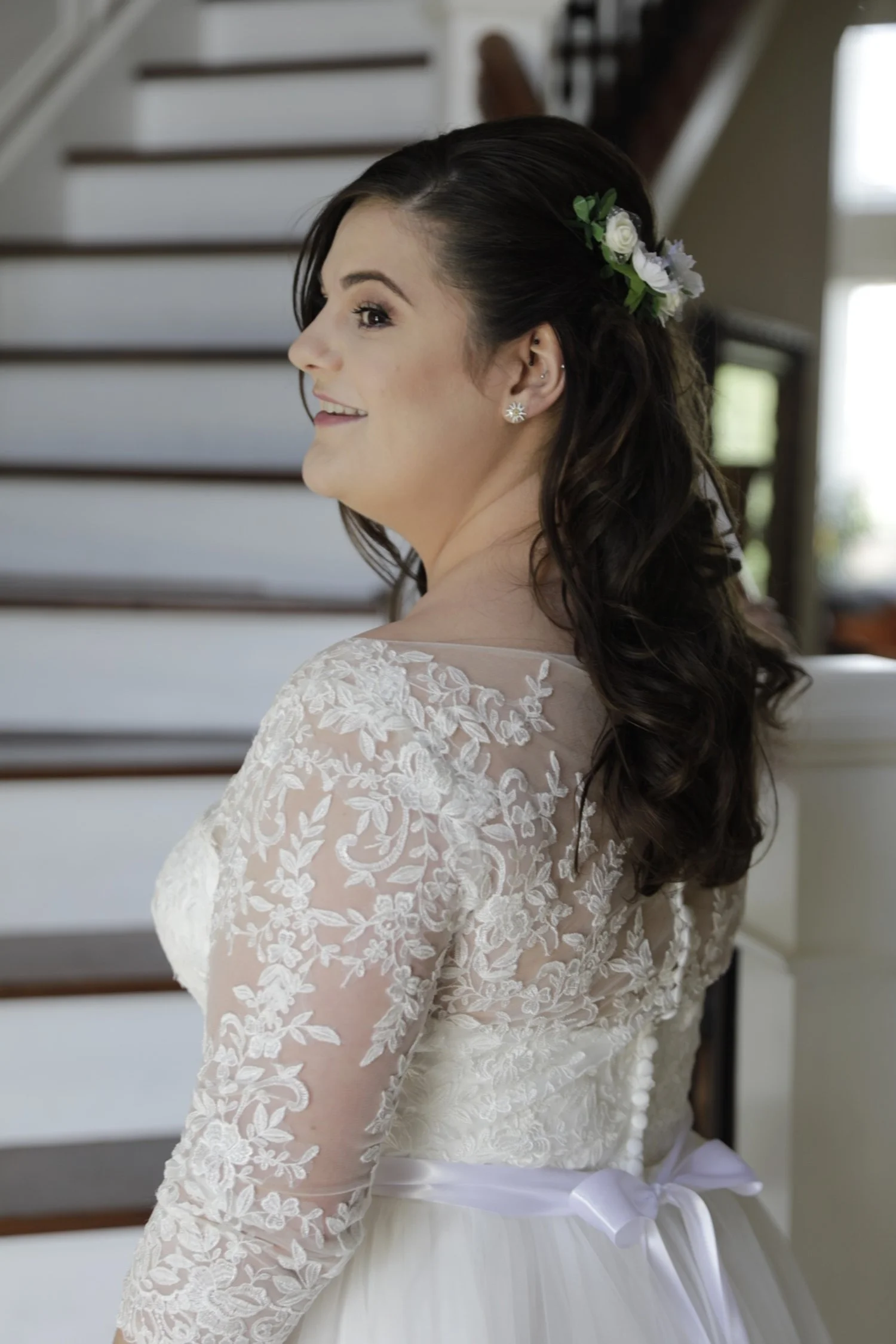 Bride in lace wedding dress with floral hair accessory, standing by staircase.