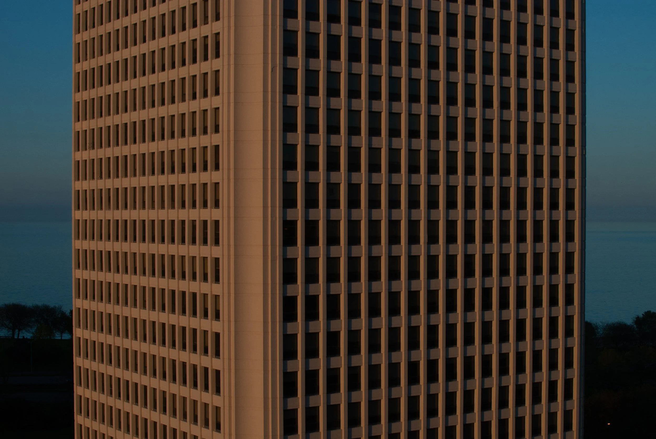 Tall building with grid-like windows against a blue sky and water backdrop.