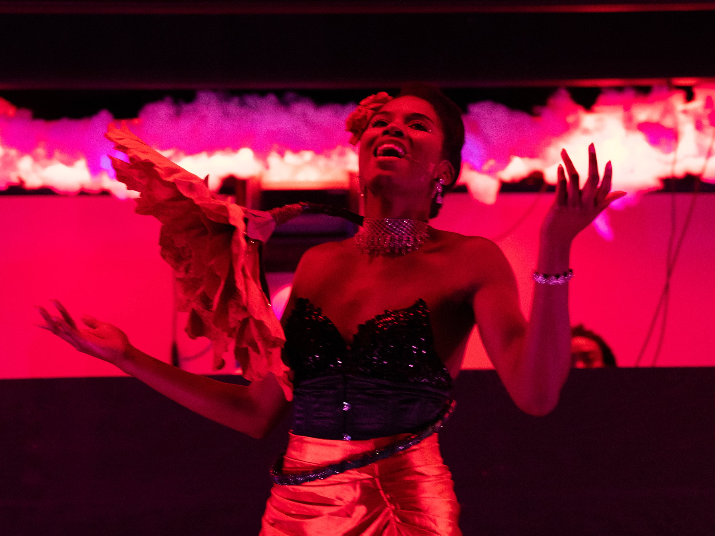 A performer in a vibrant red and black outfit on stage, with dramatic lighting. The person has their arms raised and is expressing emotion, possibly singing or dancing against a pink-lit background.