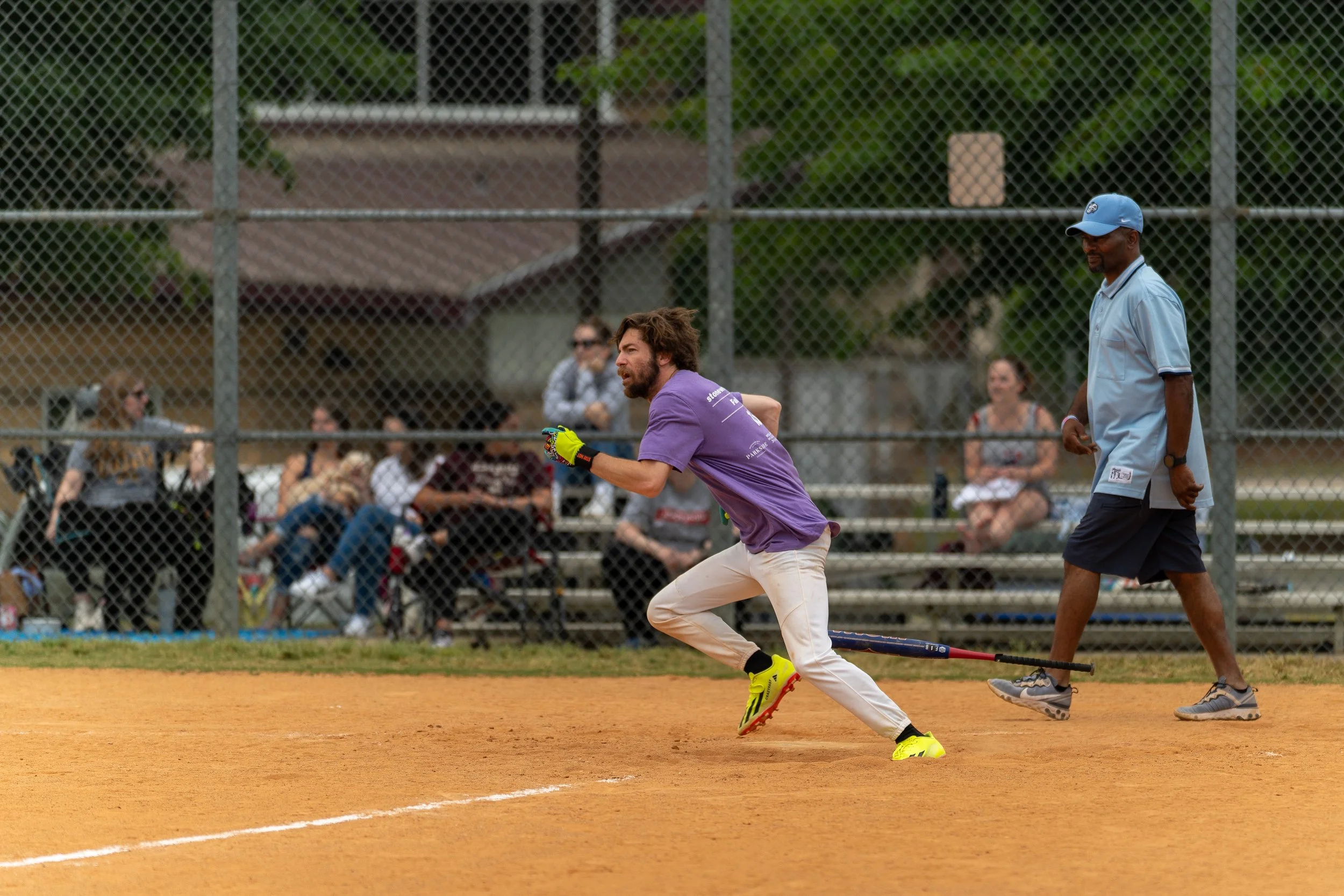 A baseball player in a purple shirt and white pants is running on a dirt field during a game. A man in a light blue shirt and cap is nearby. Spectators are seated on bleachers behind a fence in the background.