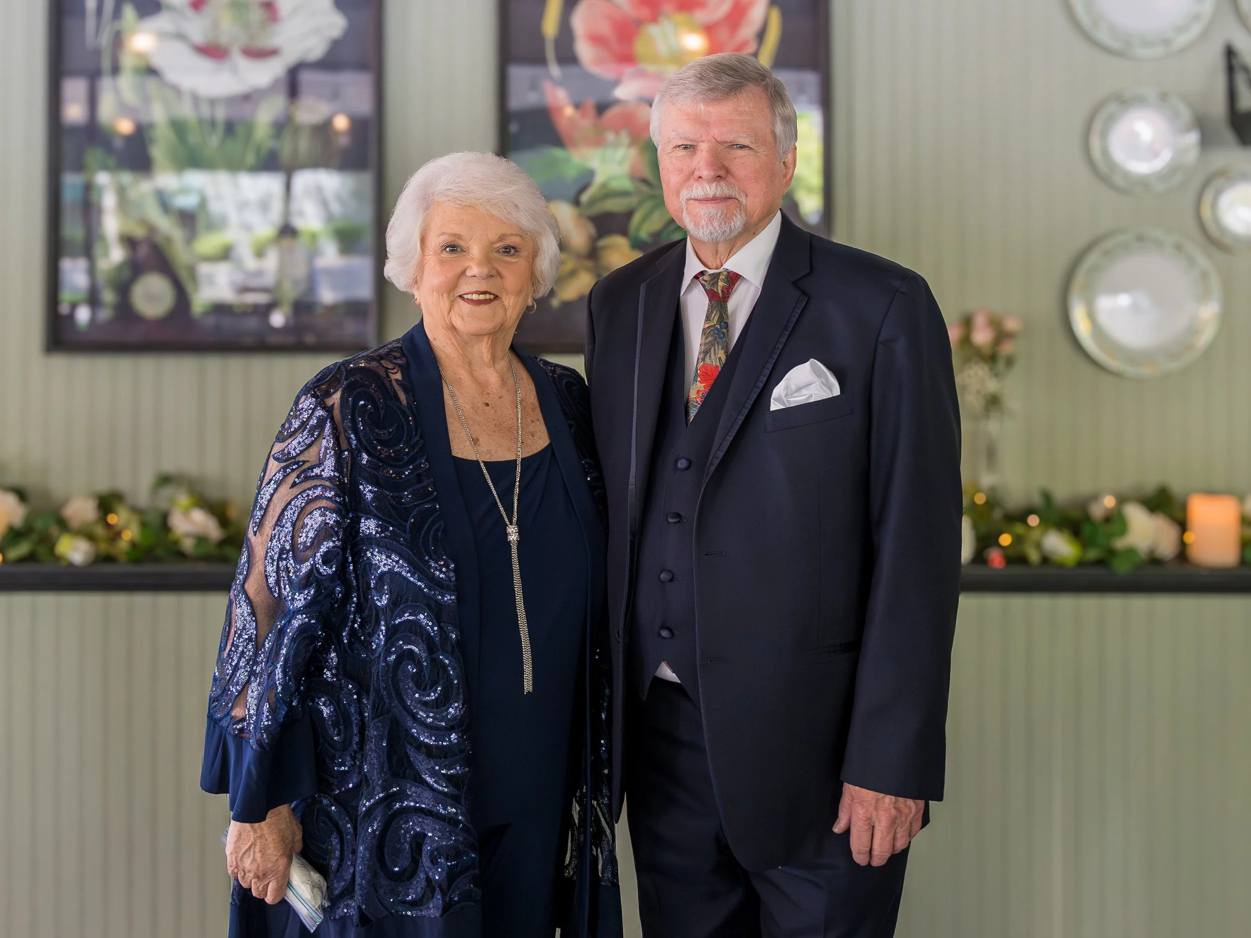 Elderly couple dressed formally, standing indoors with decorative background featuring floral artwork and tableware.