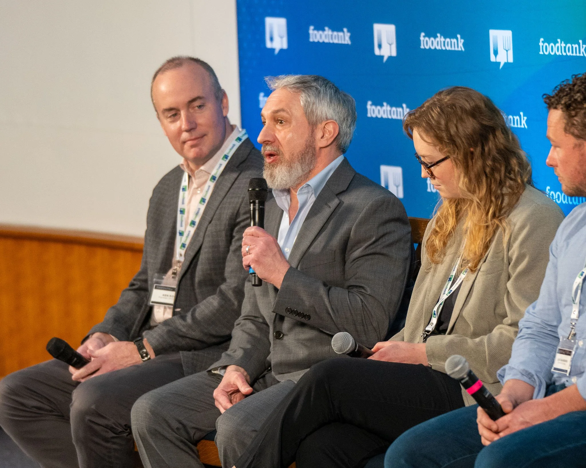 A panel discussion at a Food Tank event with three men and one woman seated, holding microphones, and engaging in conversation. The background displays the Food Tank logo.
