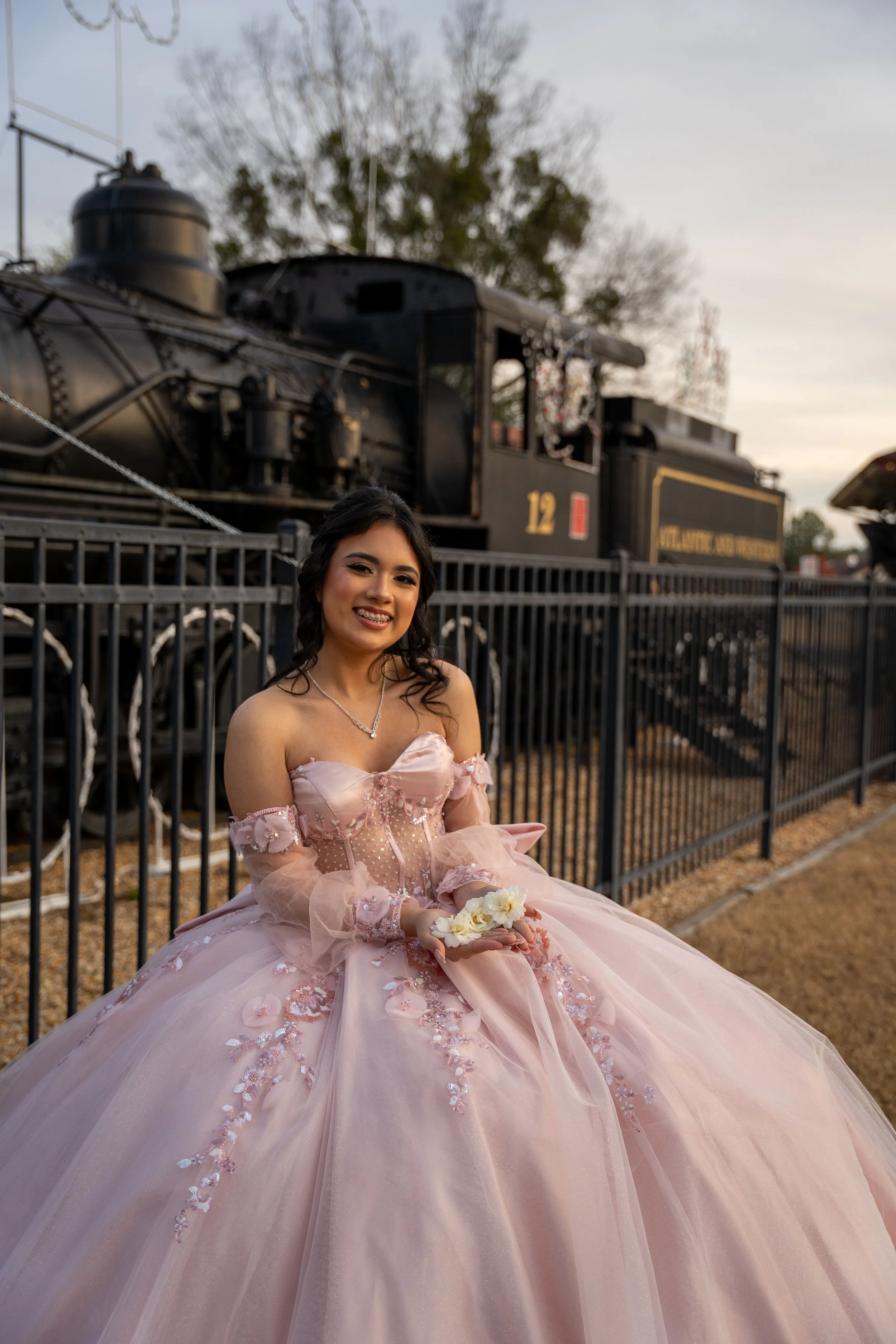 Smiling woman in a pink embellished ball gown holding white flowers beside a vintage steam locomotive.
