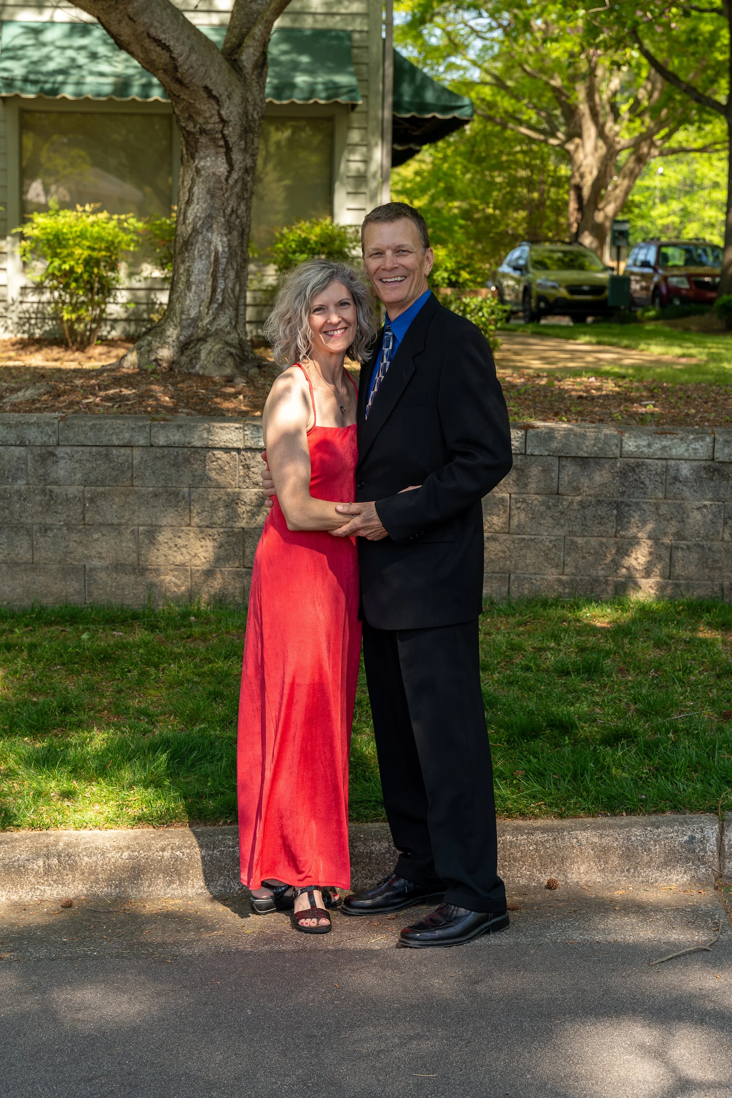 Couple posing outdoors in formal attire, man in a black suit and woman in a red dress, standing on a paved path with greenery in the background.