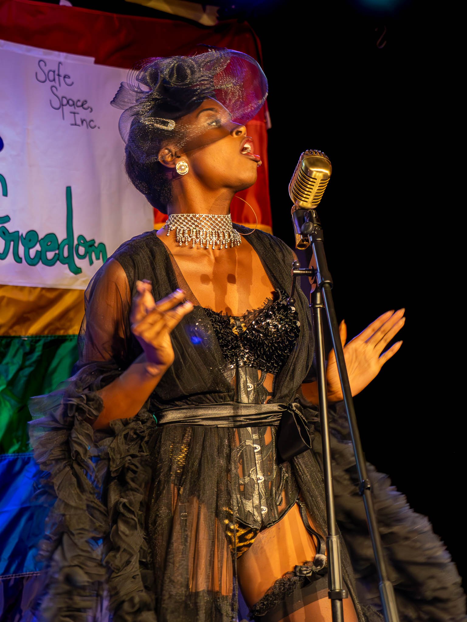 Performer in elaborate costume and hat singing into a vintage microphone on stage, with colorful backdrop and "Safe Space, Inc. Freedom" banner visible.