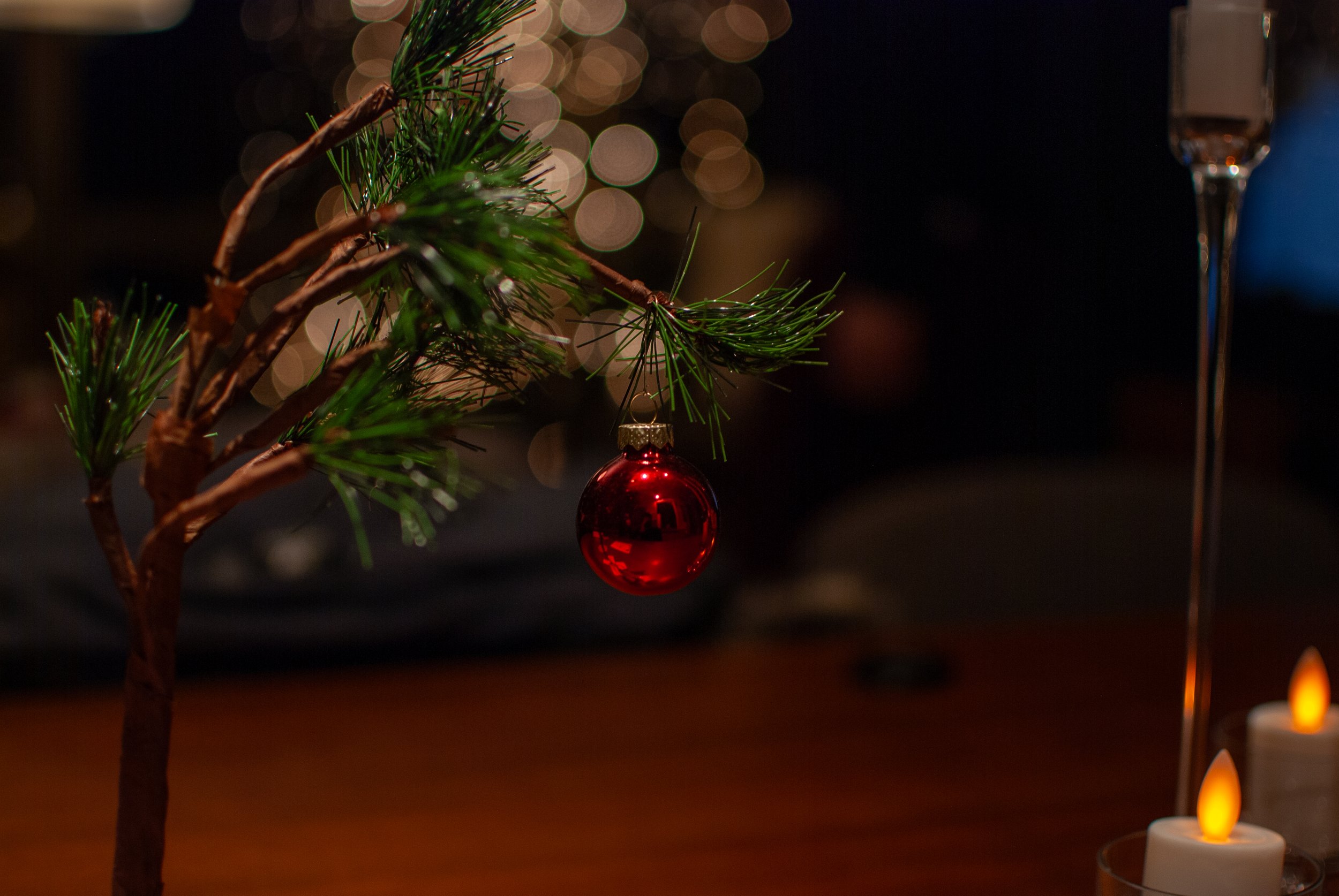 Small artificial Christmas tree with a red ornament and candles on a table, blurred background with bokeh lights.