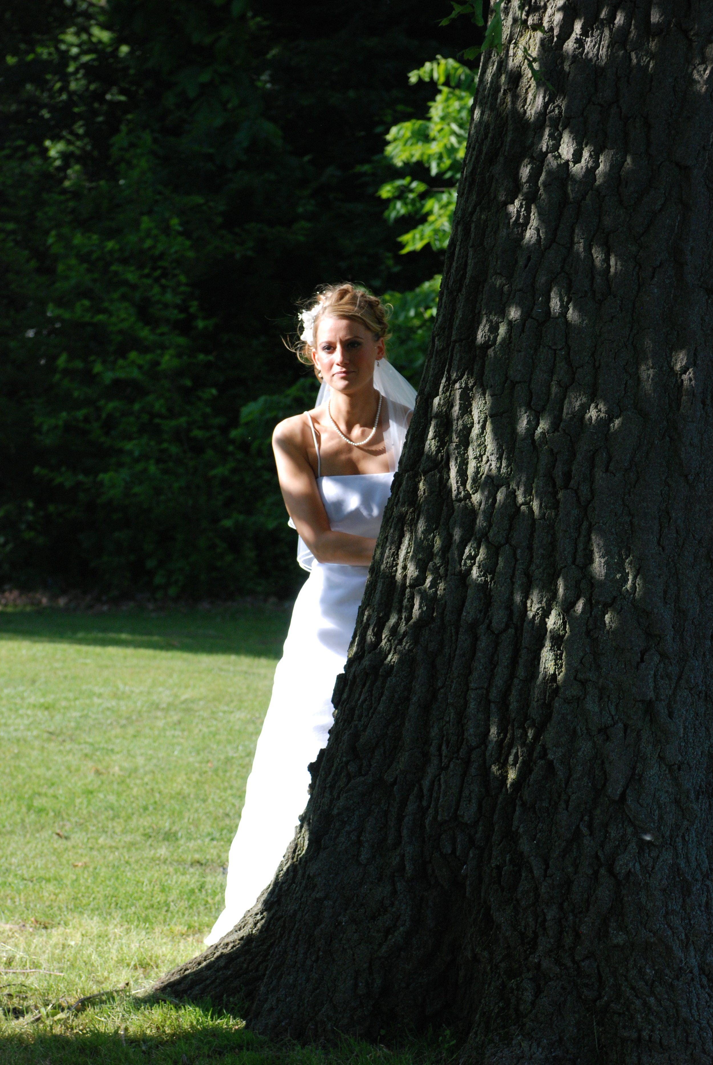 Bride in white dress peeking from behind a tree in a park.