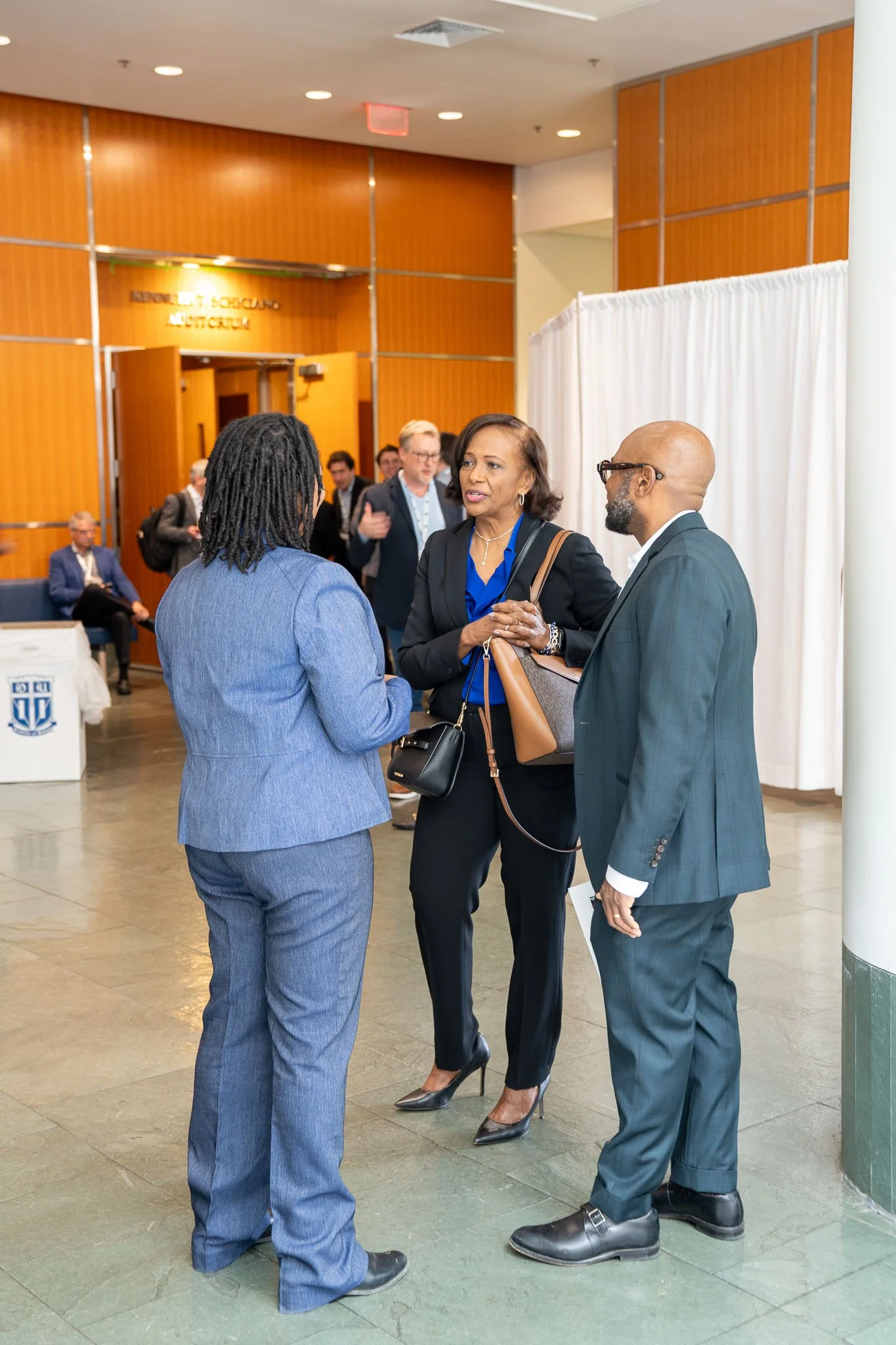 Three people in formal attire talking in a hallway with wood paneling and a white curtain. A sign for Kenneth L. Schilling Auditorium is visible in the background.