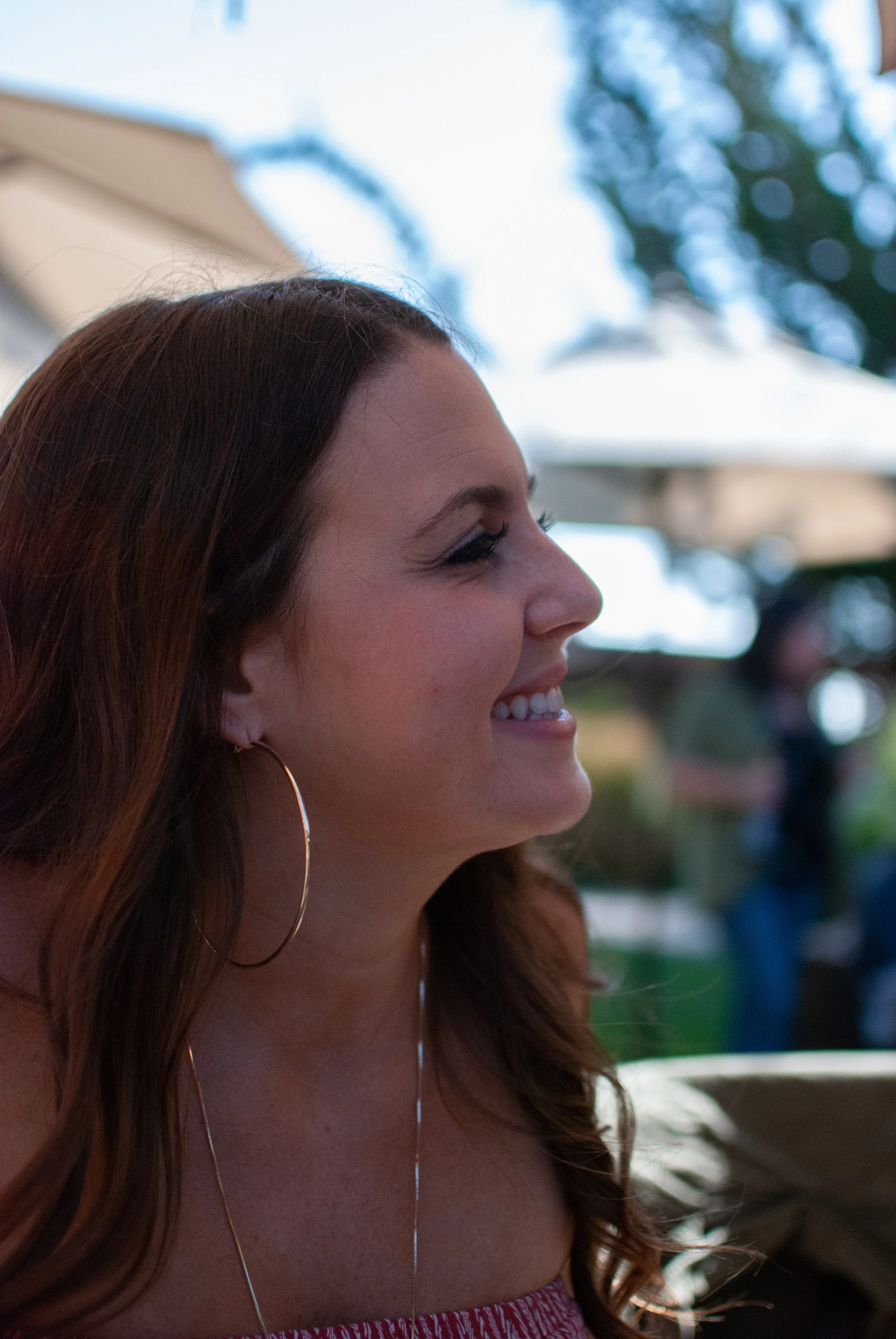Side profile of a smiling woman with long brown hair, wearing large hoop earrings, outdoors.