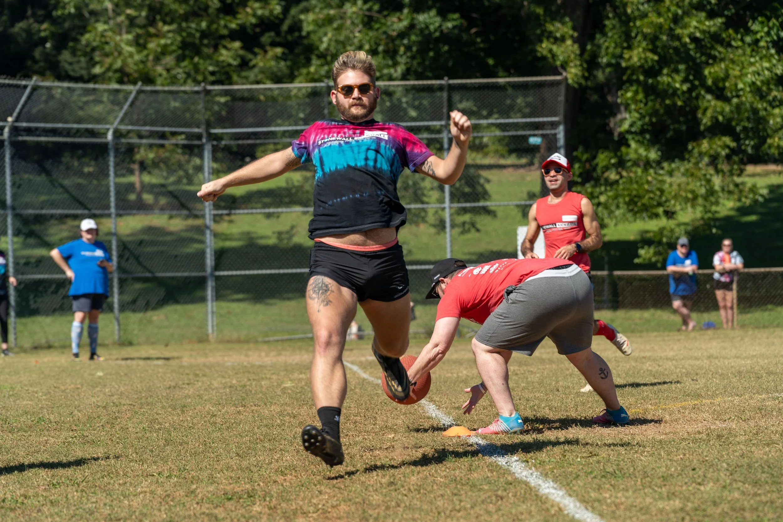 A group of people playing kickball on a grassy field. A player in a colorful shirt and shorts kicks the ball, while another in red prepares to catch it. Spectators and a fence are visible in the background.