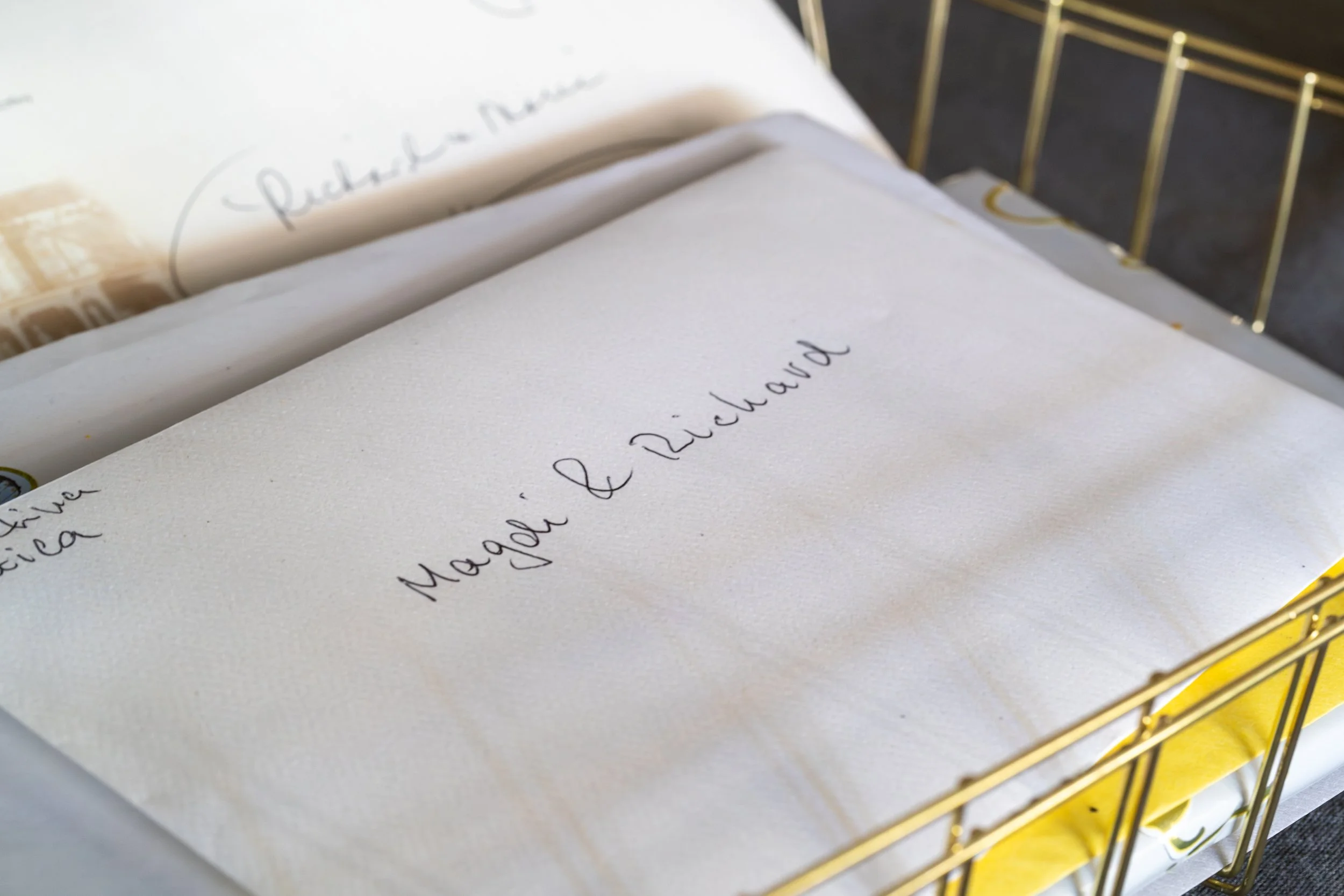 A close-up of a stack of letters in a wire basket, the top envelope labeled "Maggi & Richard."