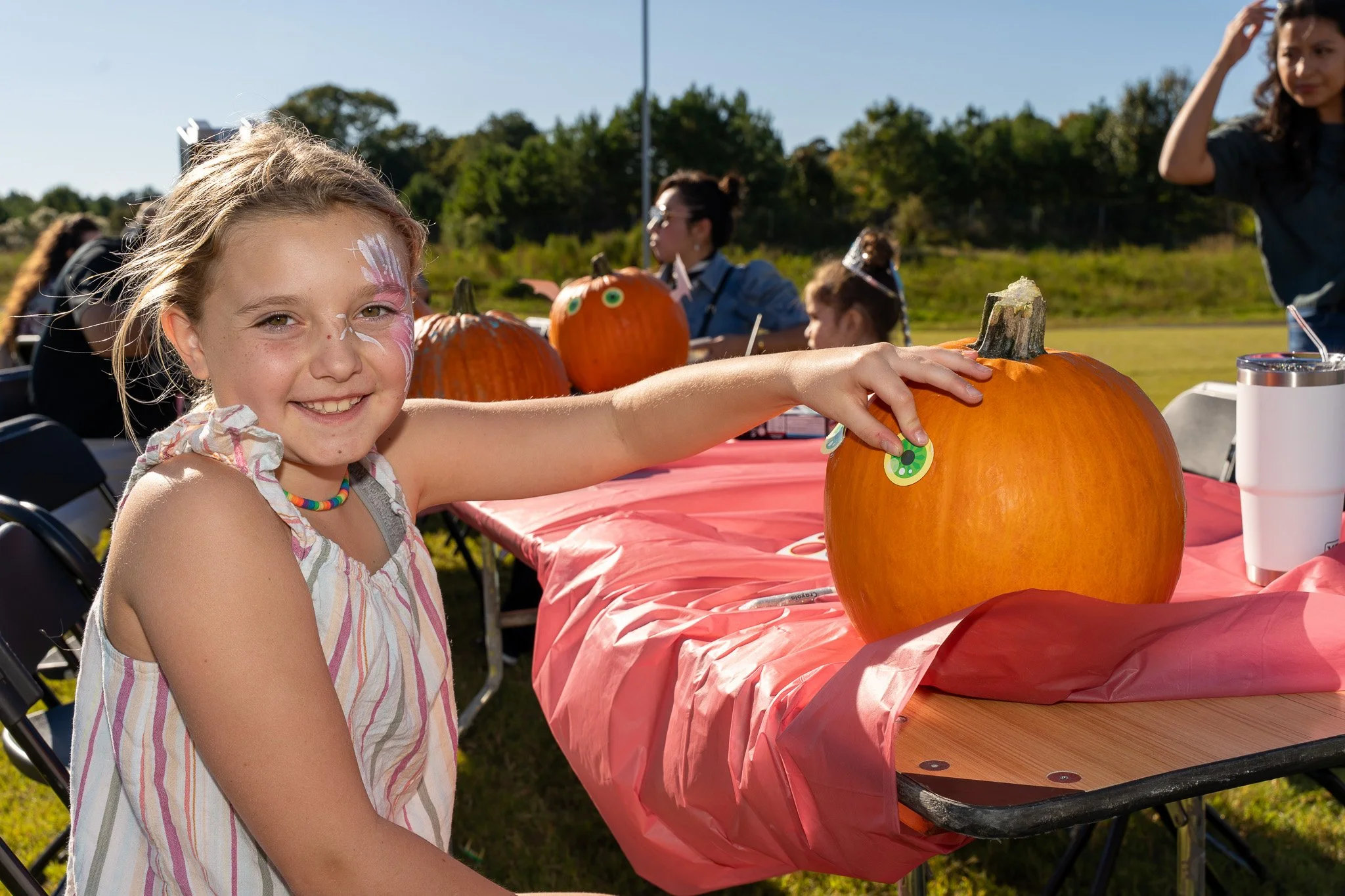 Smiling girl with butterfly face paint decorating a large pumpkin at an outdoor fall festival