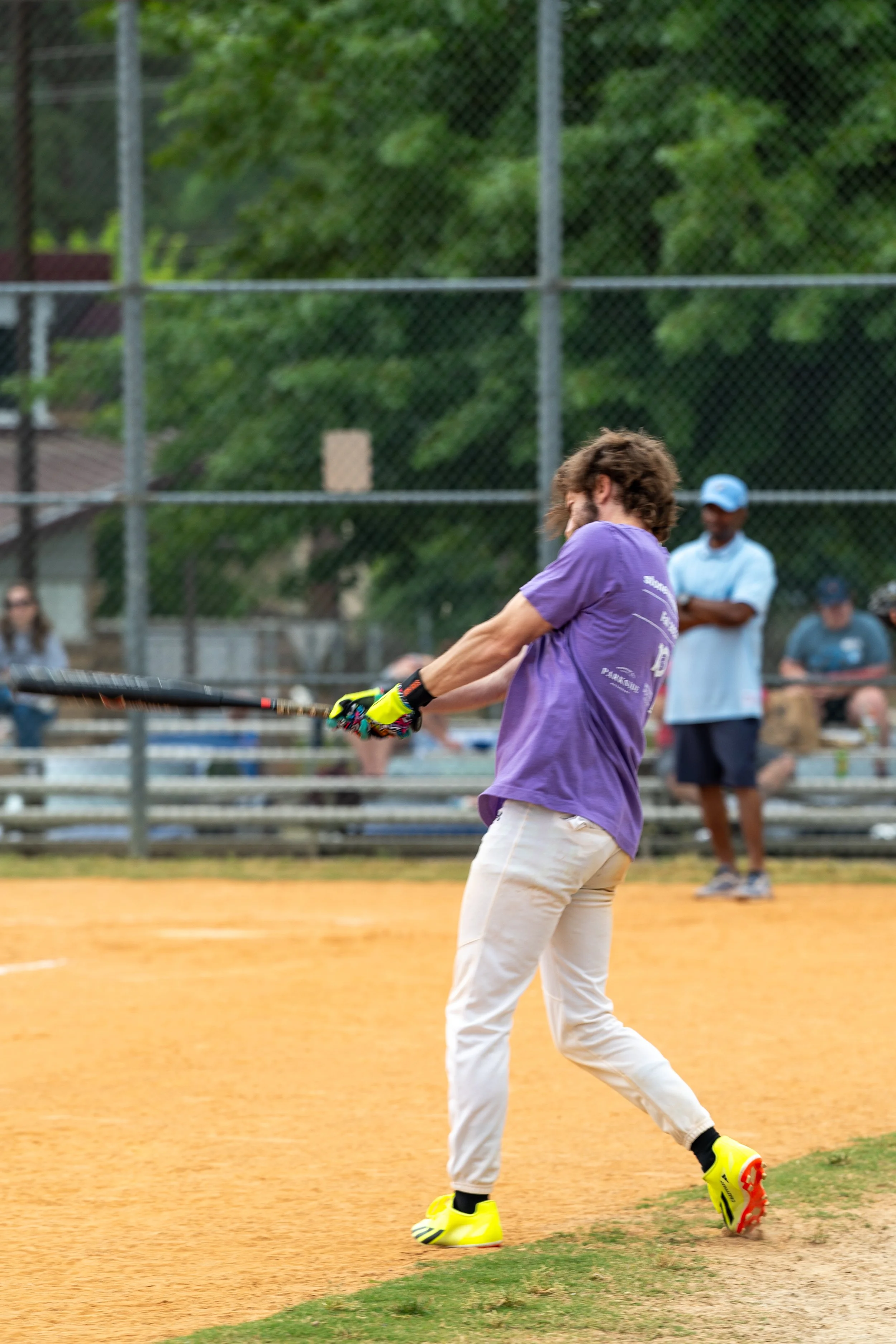 A person wearing a purple shirt and white pants swinging a baseball bat on a dirt field, with a chain-link fence and some spectators in the background.