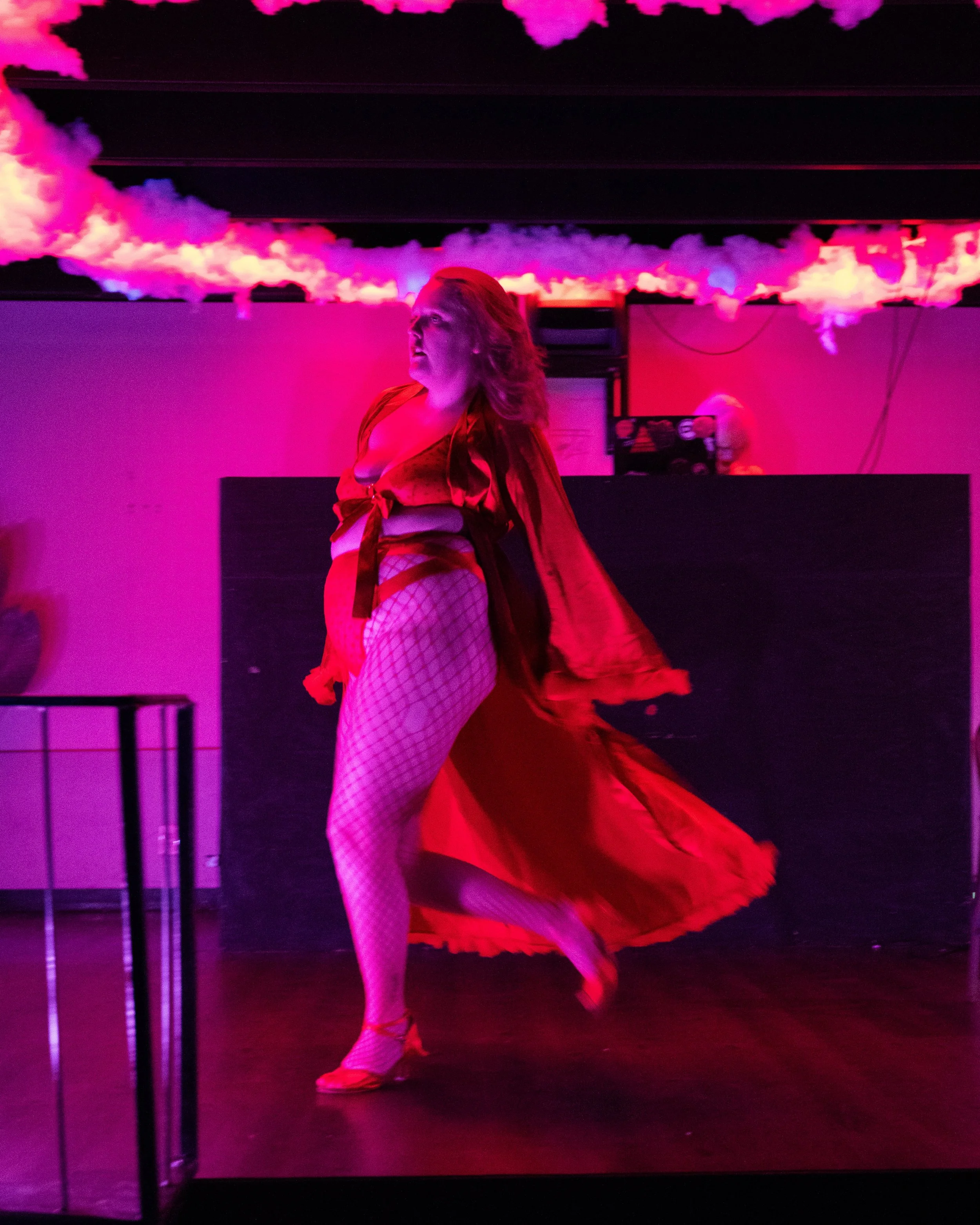 Person performing on stage with dramatic lighting and cloud-like decorations overhead, wearing a red outfit and fishnet stockings.