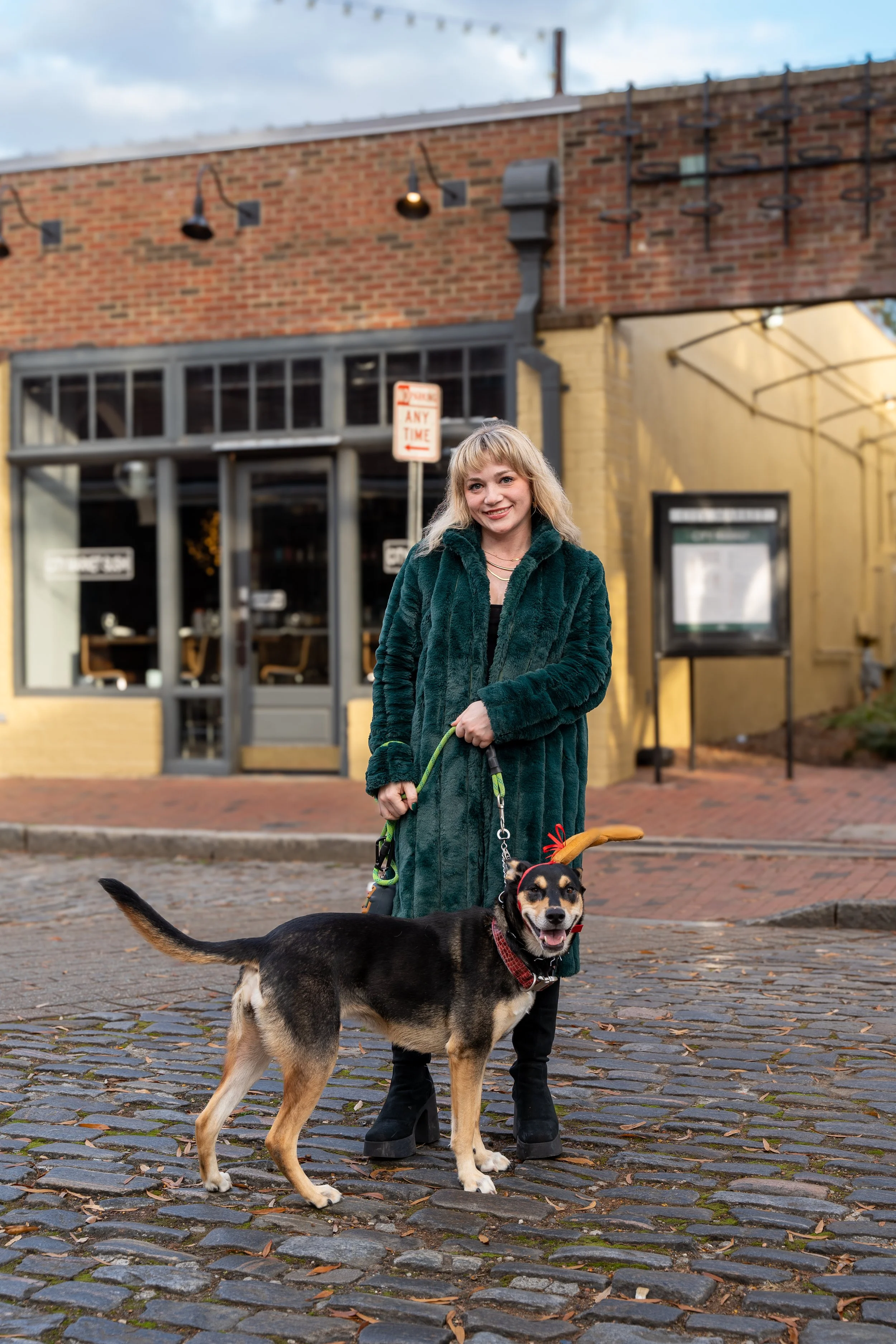 Woman wearing a green coat standing on cobblestone street with a dog wearing a red bandana and reindeer antlers, in front of a brick building.