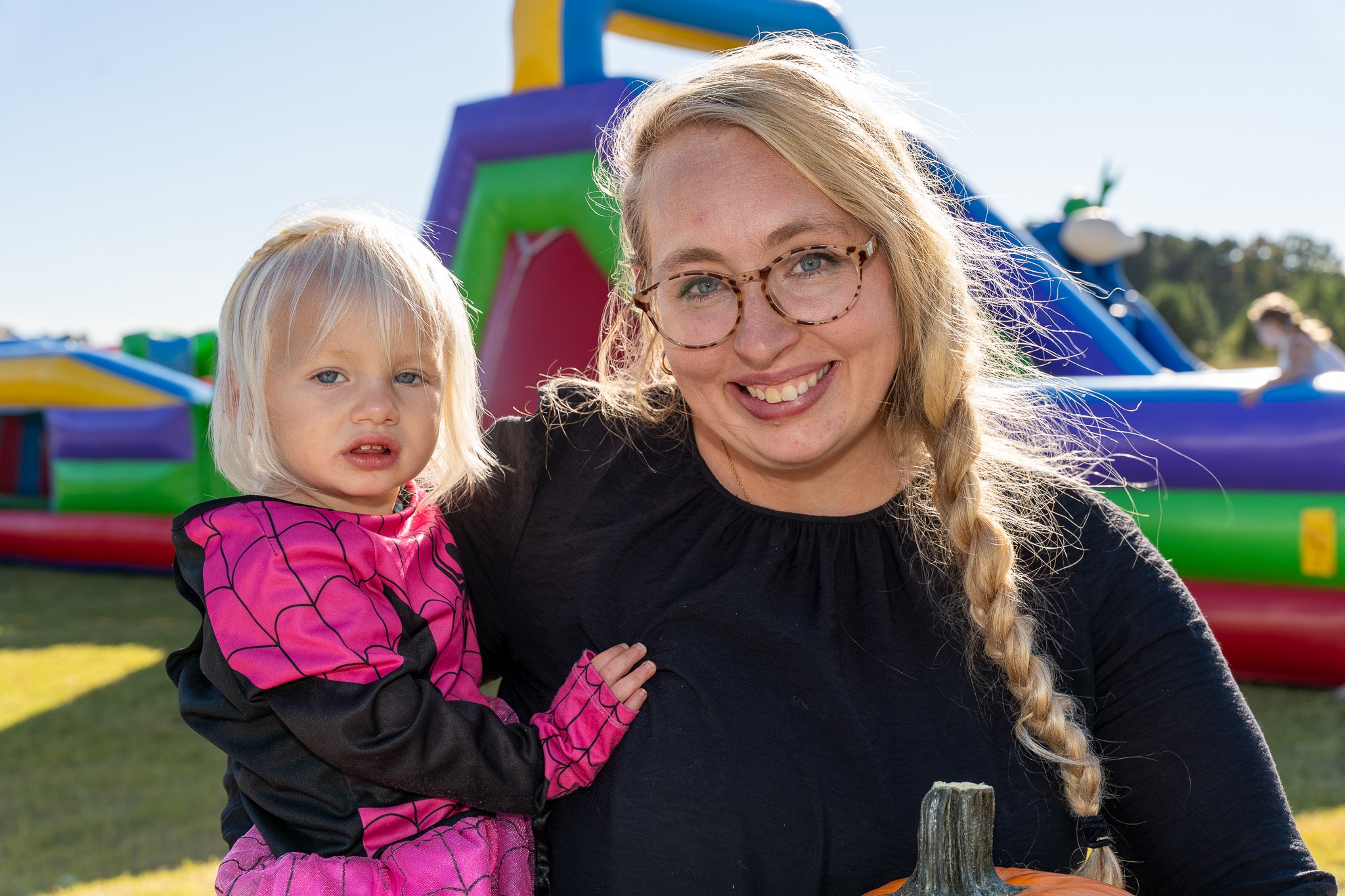 Smiling mother holding a young child in a pink Spider-Man costume at an outdoor fall festival with an inflatable bounce house