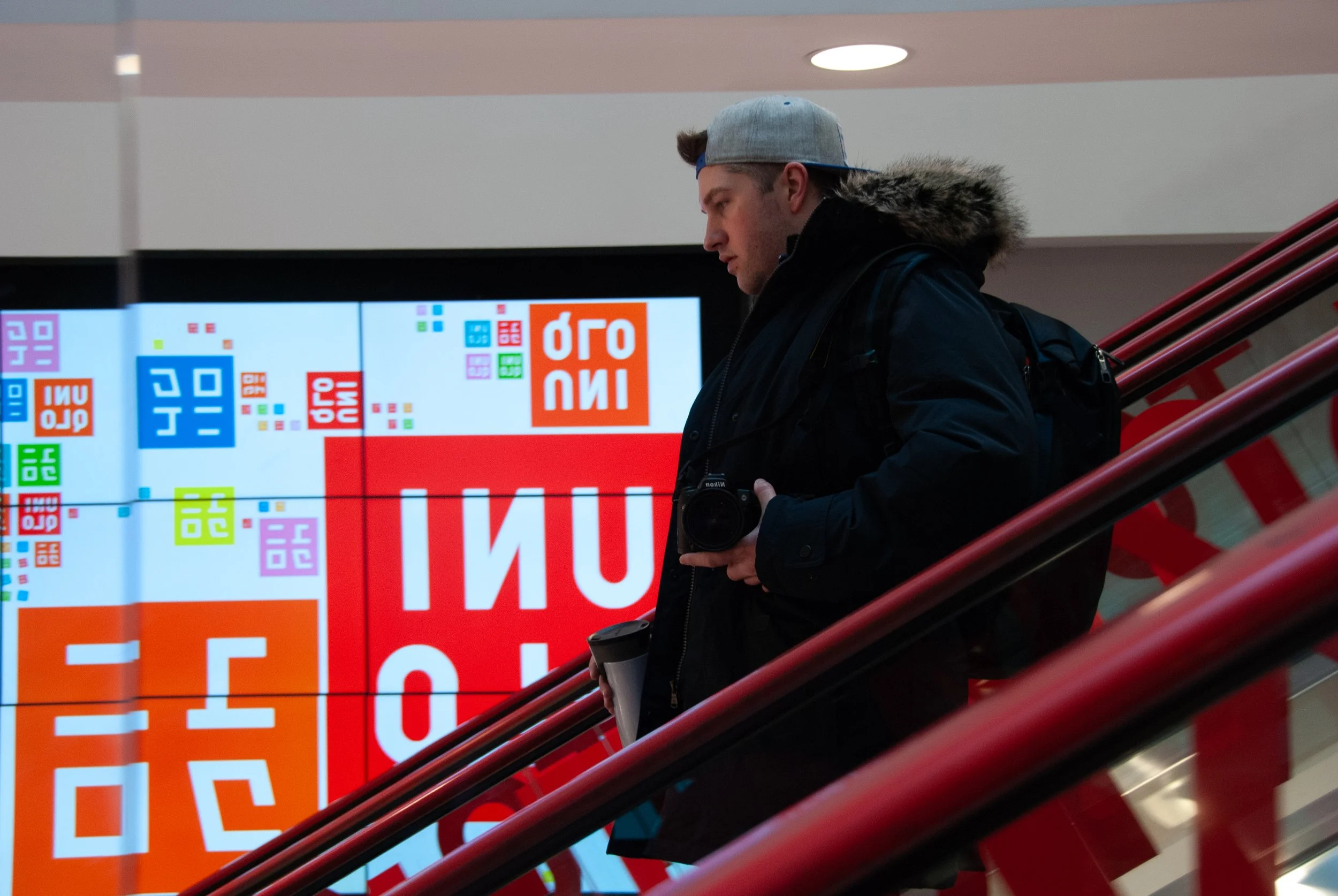 Person with a camera and coffee cup on an escalator in front of colorful digital signs.