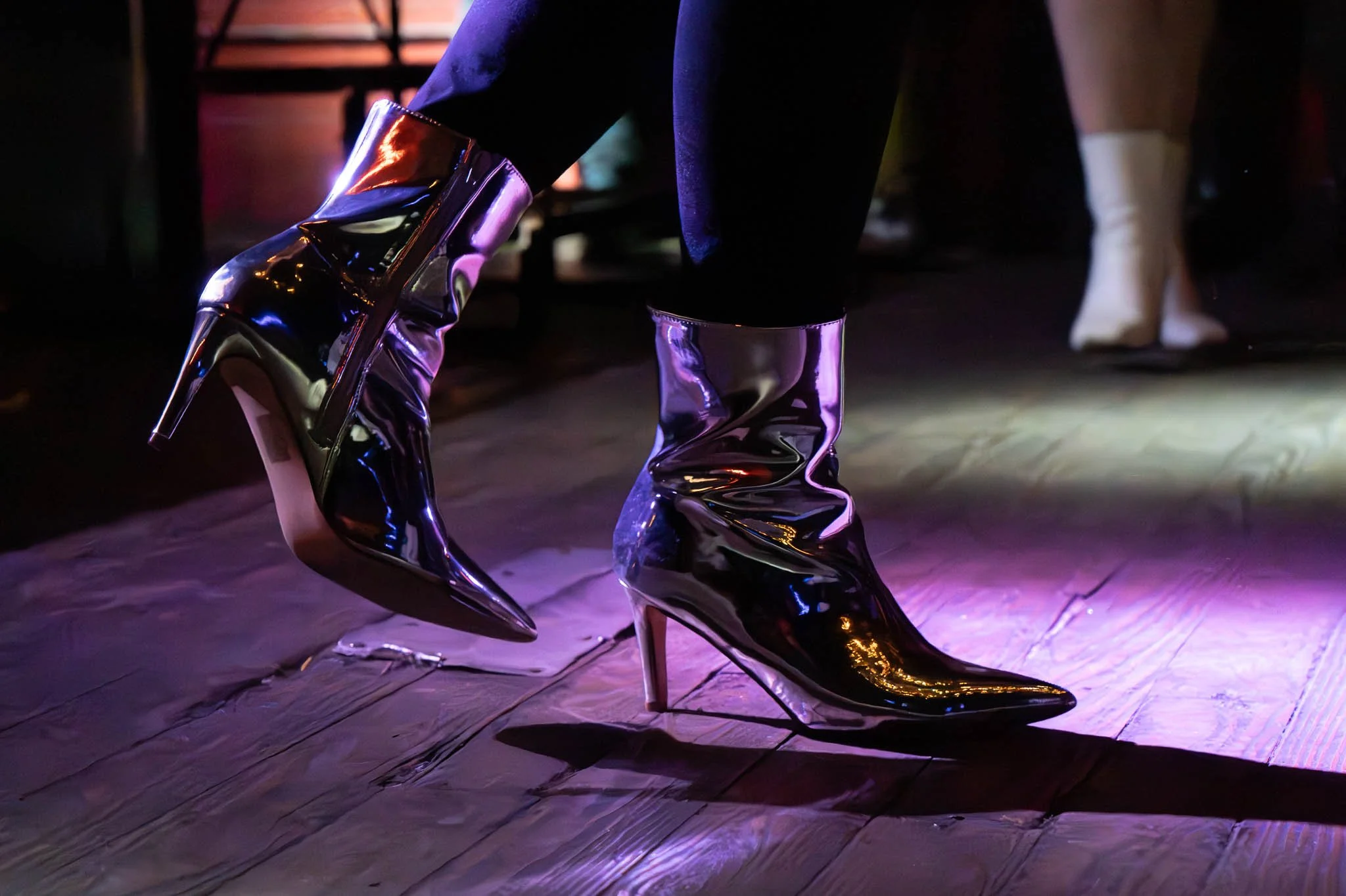 Close-up of metallic silver high-heeled boots on a wooden floor with colorful lighting.