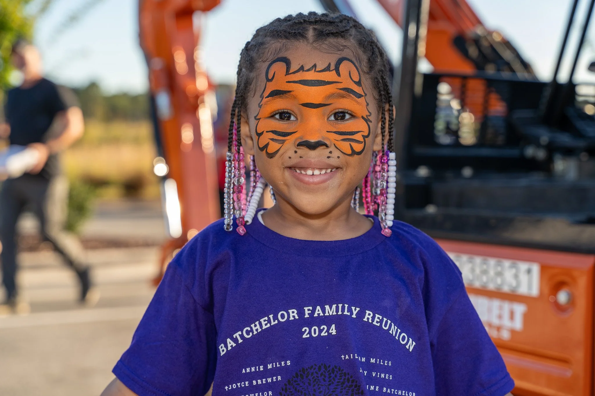 Smiling child with braided, beaded hair and orange tiger face paint standing near an orange fire truck.