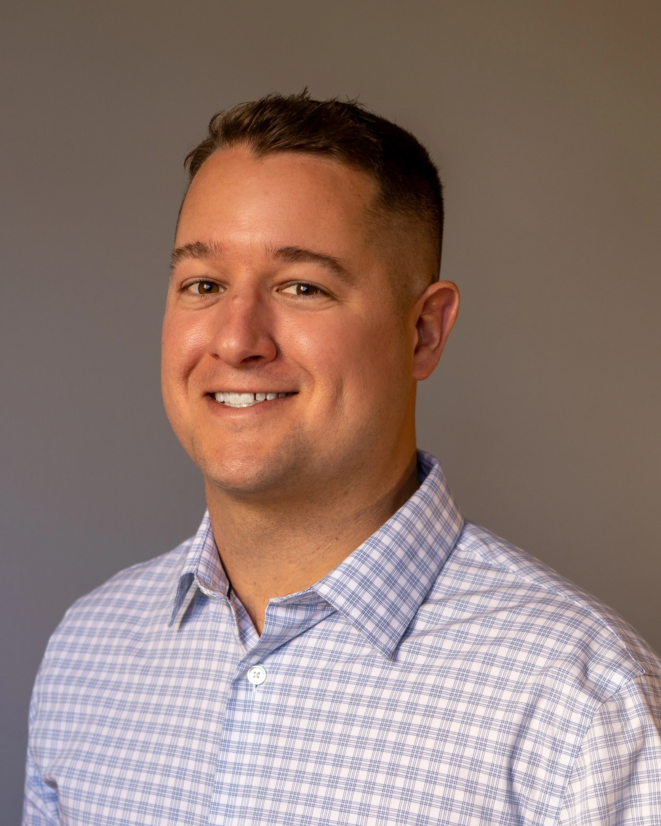 A man wearing a checkered shirt smiles against a neutral background.
