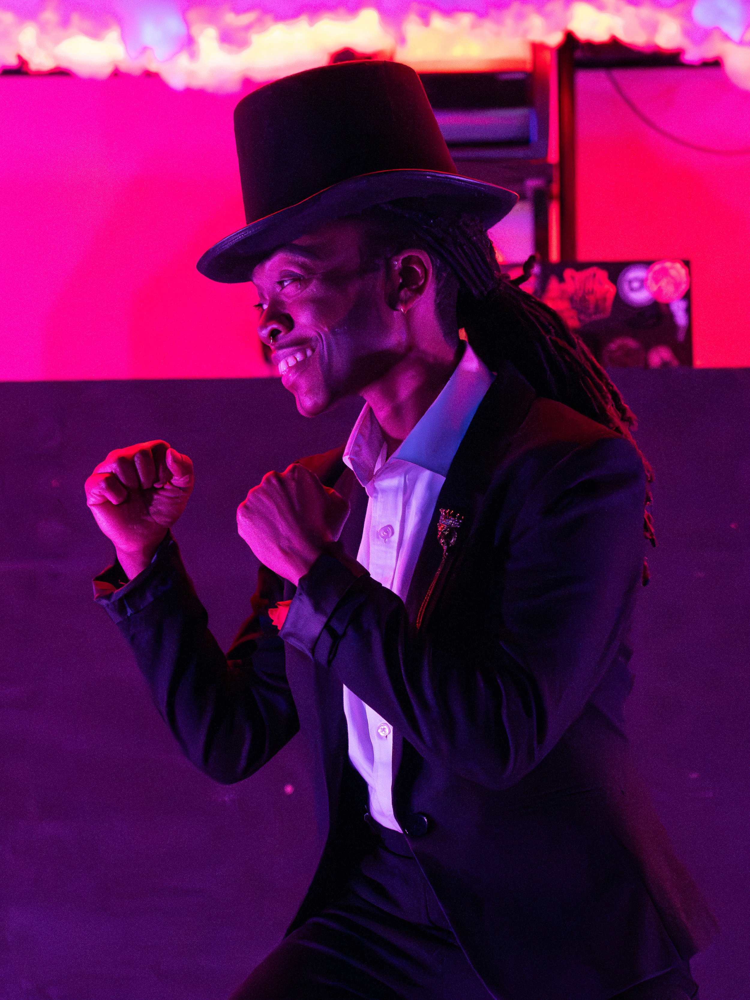 A person in a top hat and suit, posing with fists up, in a brightly lit environment with purple and pink lighting.