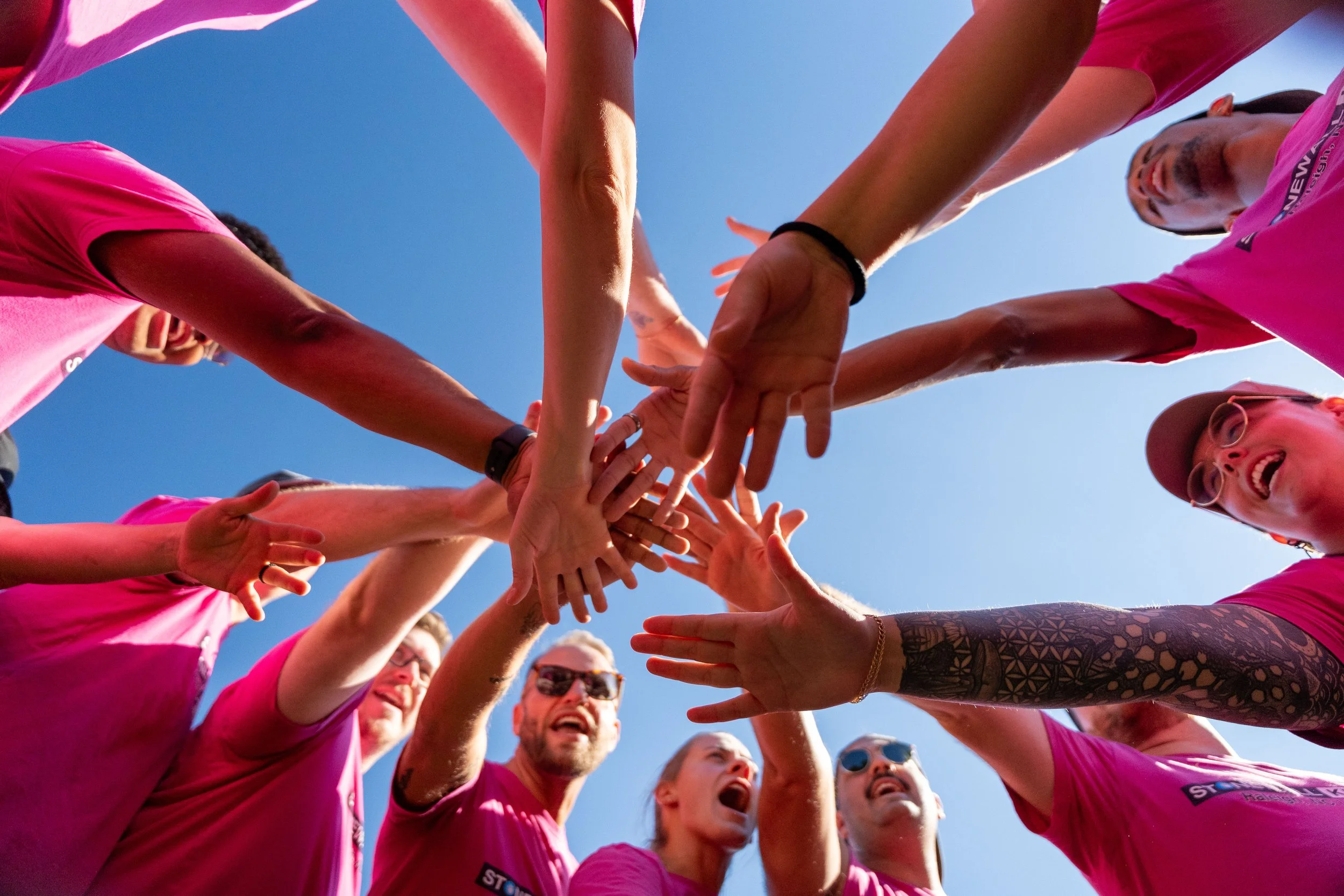Group of people in pink shirts with hands joined in a circle against a blue sky.