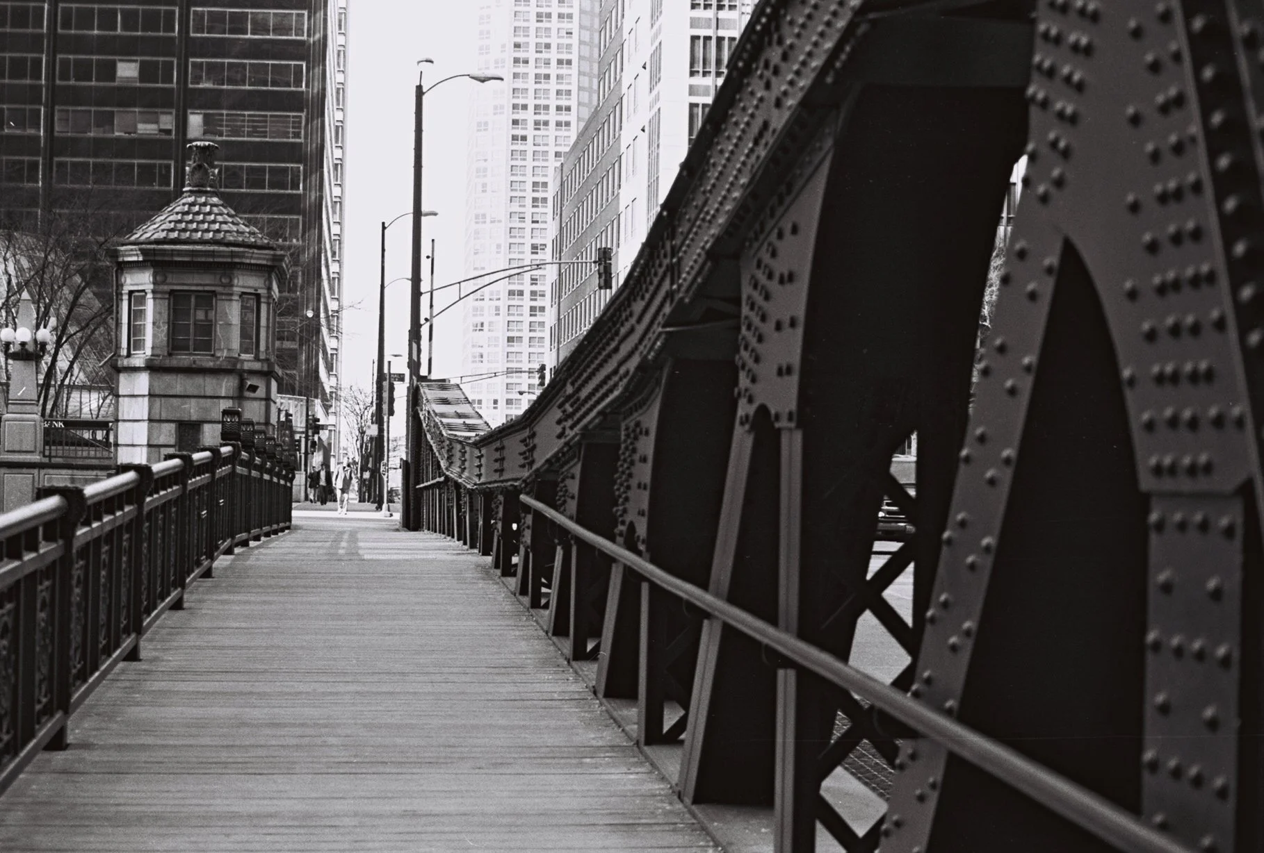 Black and white photo of an urban bridge with metal beams, a pedestrian walkway, and city buildings in the background.