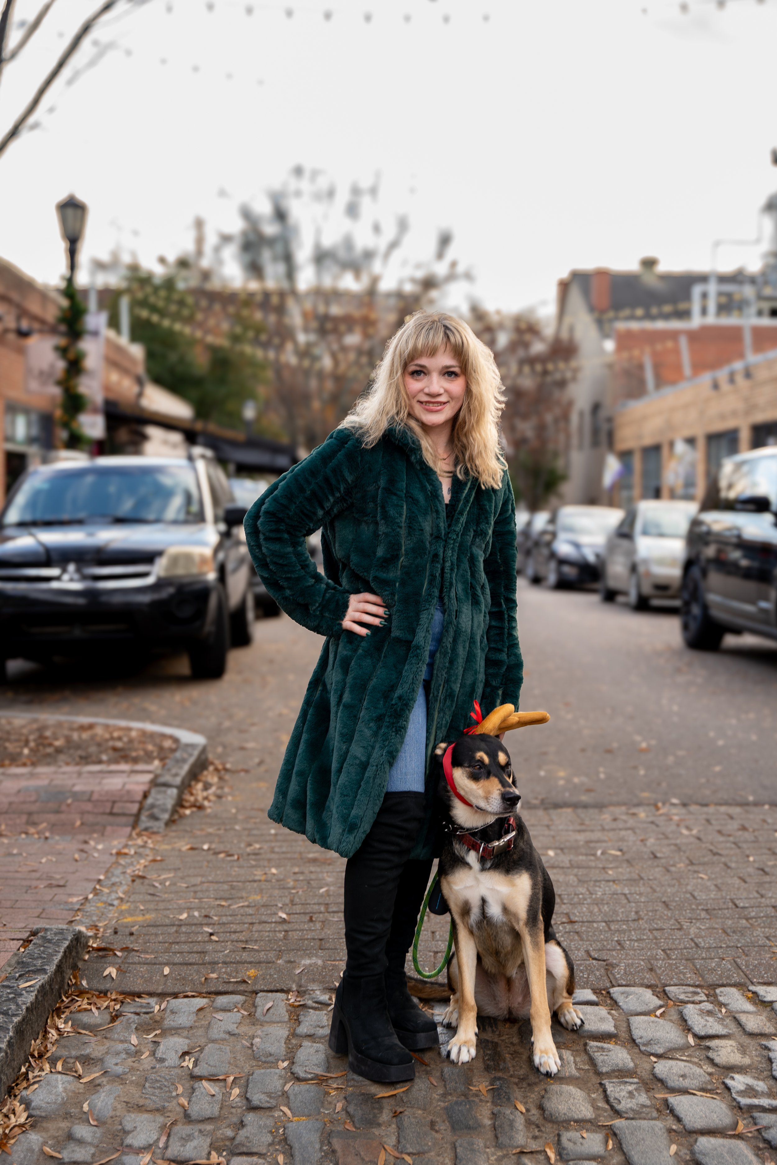 Woman in a green fur coat standing on a cobblestone street with a dog wearing reindeer antlers.