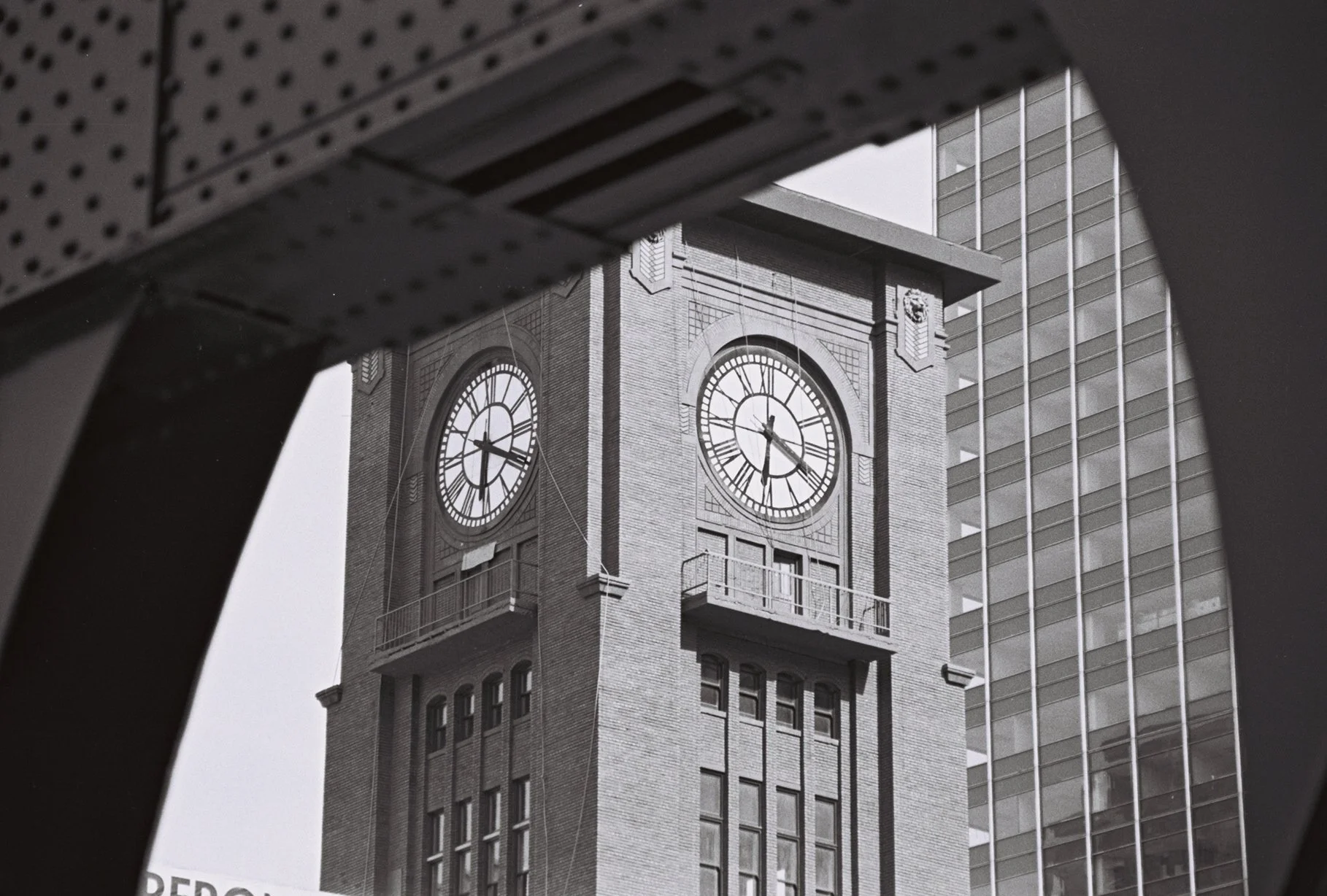 Black and white photo of a clock tower with two clock faces framed by structural elements of a bridge. The tower is made of brick and adjacent to a modern glass building.