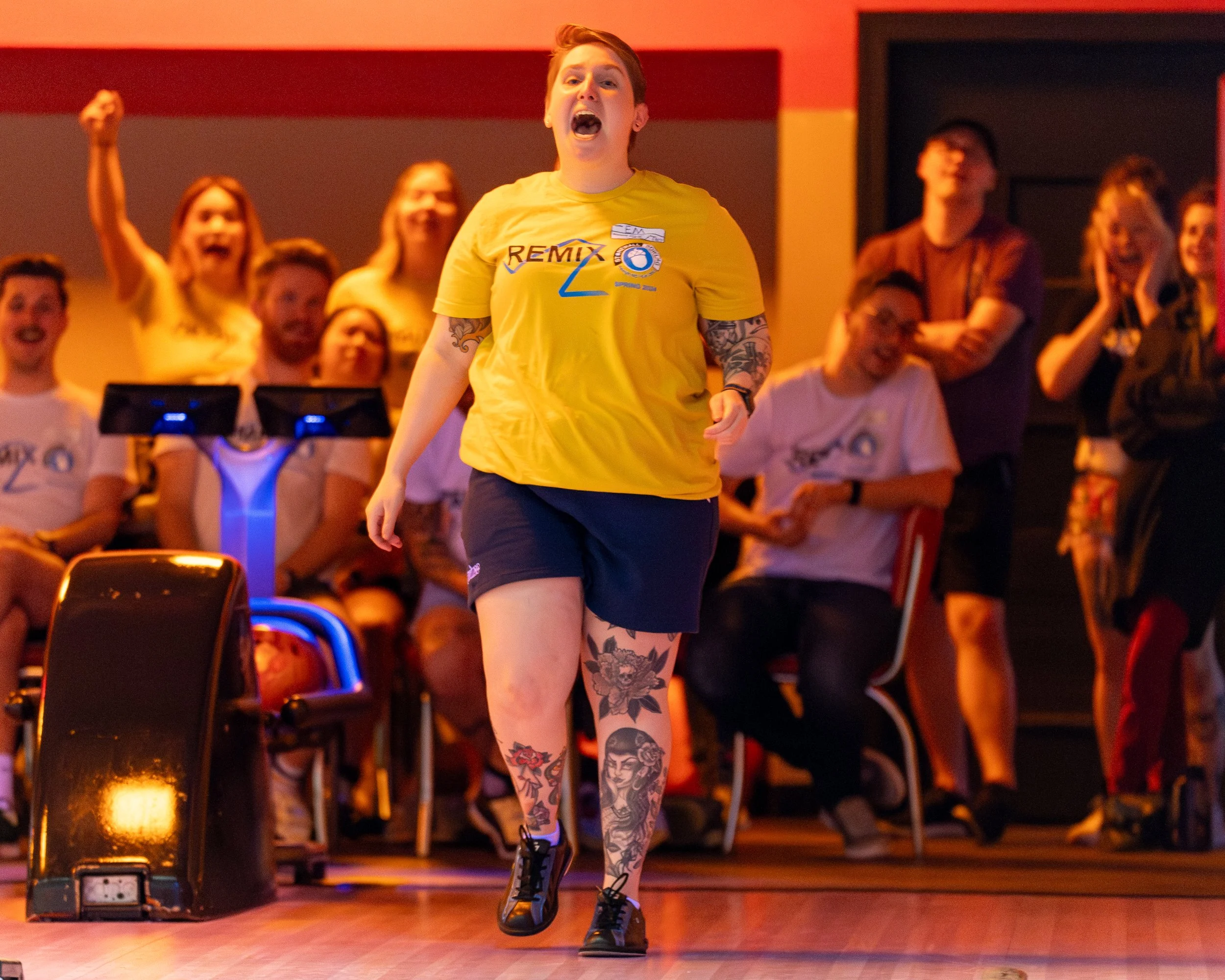 A person in a yellow shirt and shorts with tattoos is excitedly walking down a bowling lane in front of an audience. The crowd in the background is cheering and clapping.