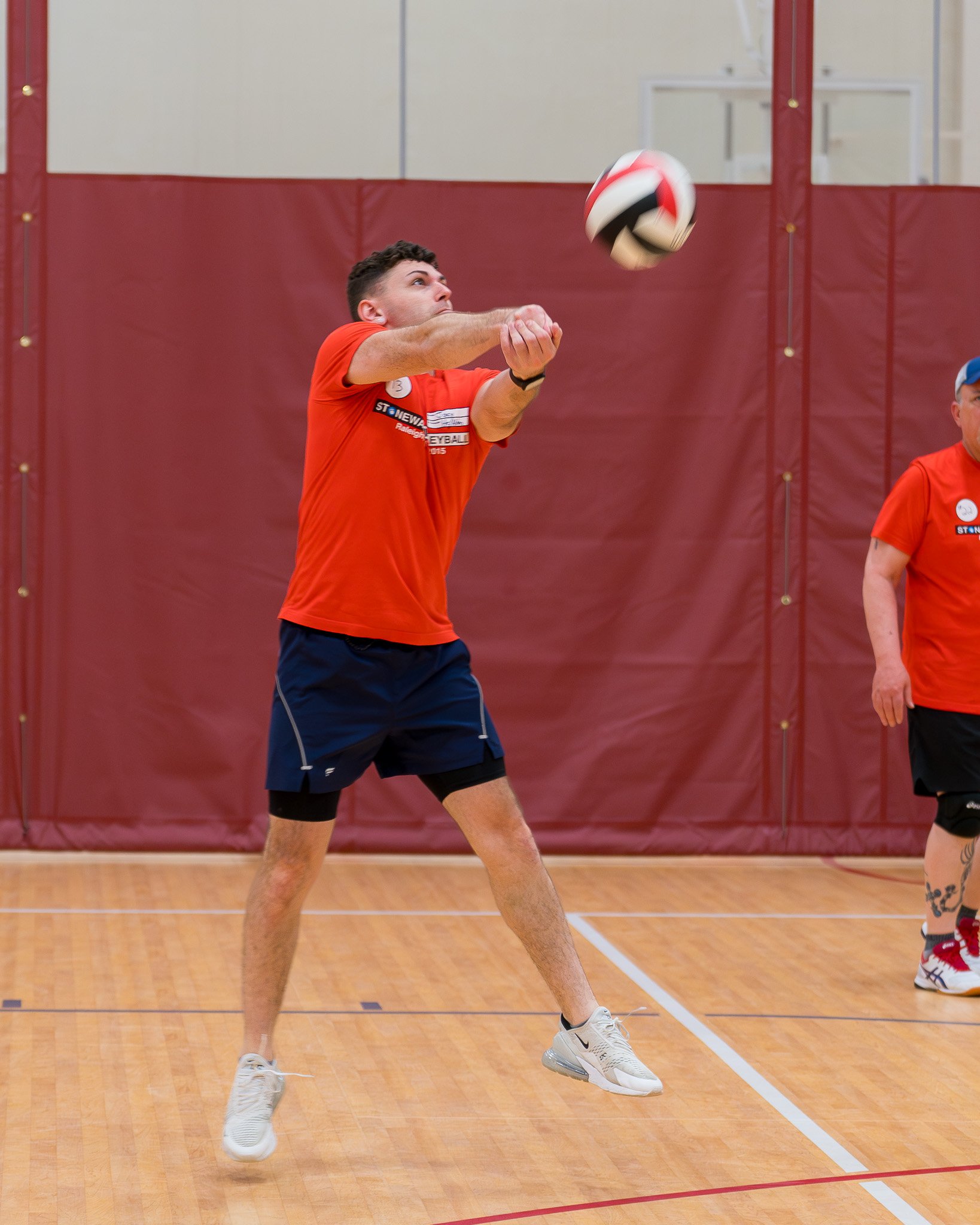 A man in an orange shirt and blue shorts playing volleyball indoors, jumping to hit the ball with both hands. Another person in an orange shirt is partially visible on the right.
