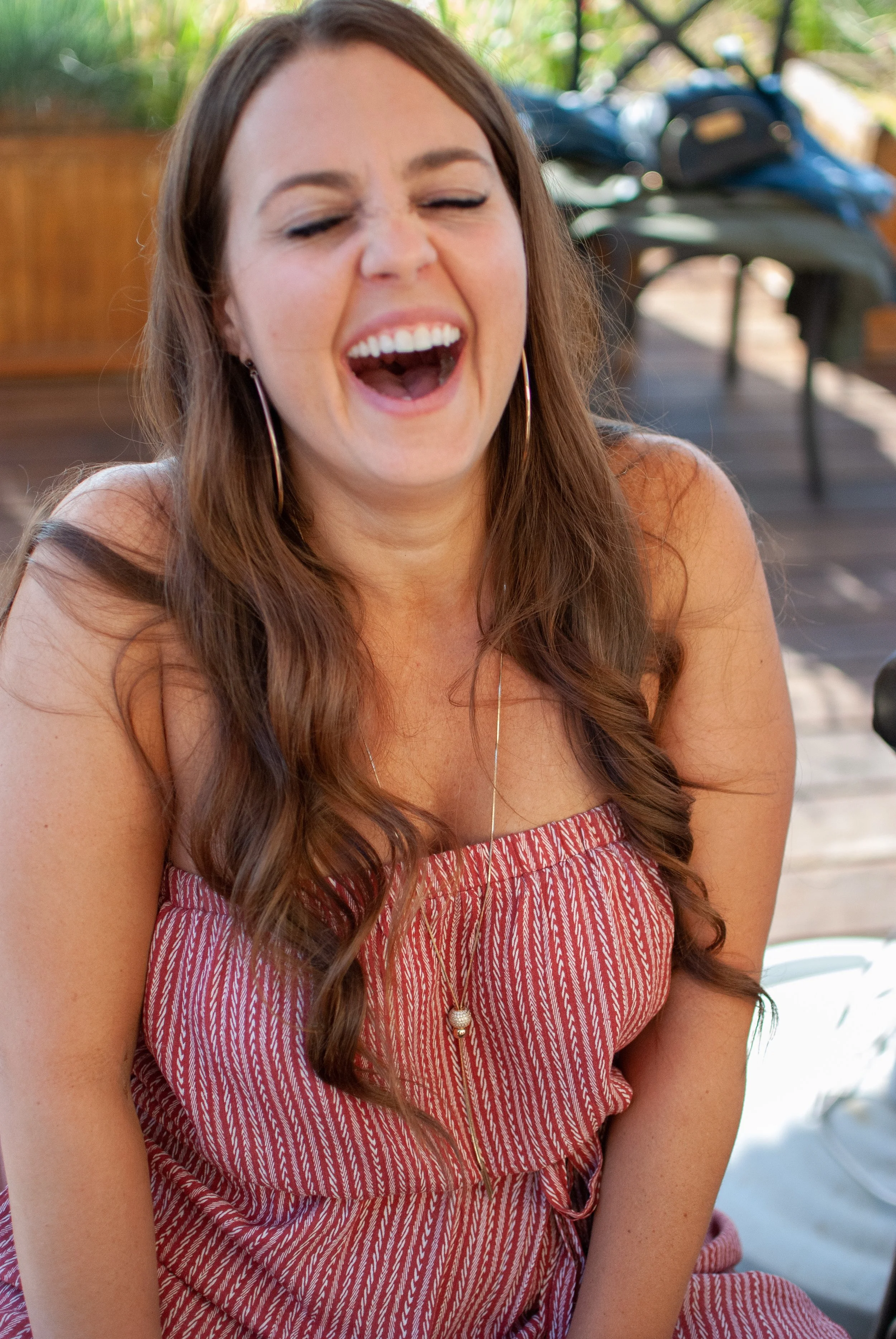 A woman with long brown hair laughing joyfully, wearing a red and white striped dress, seated outdoors.
