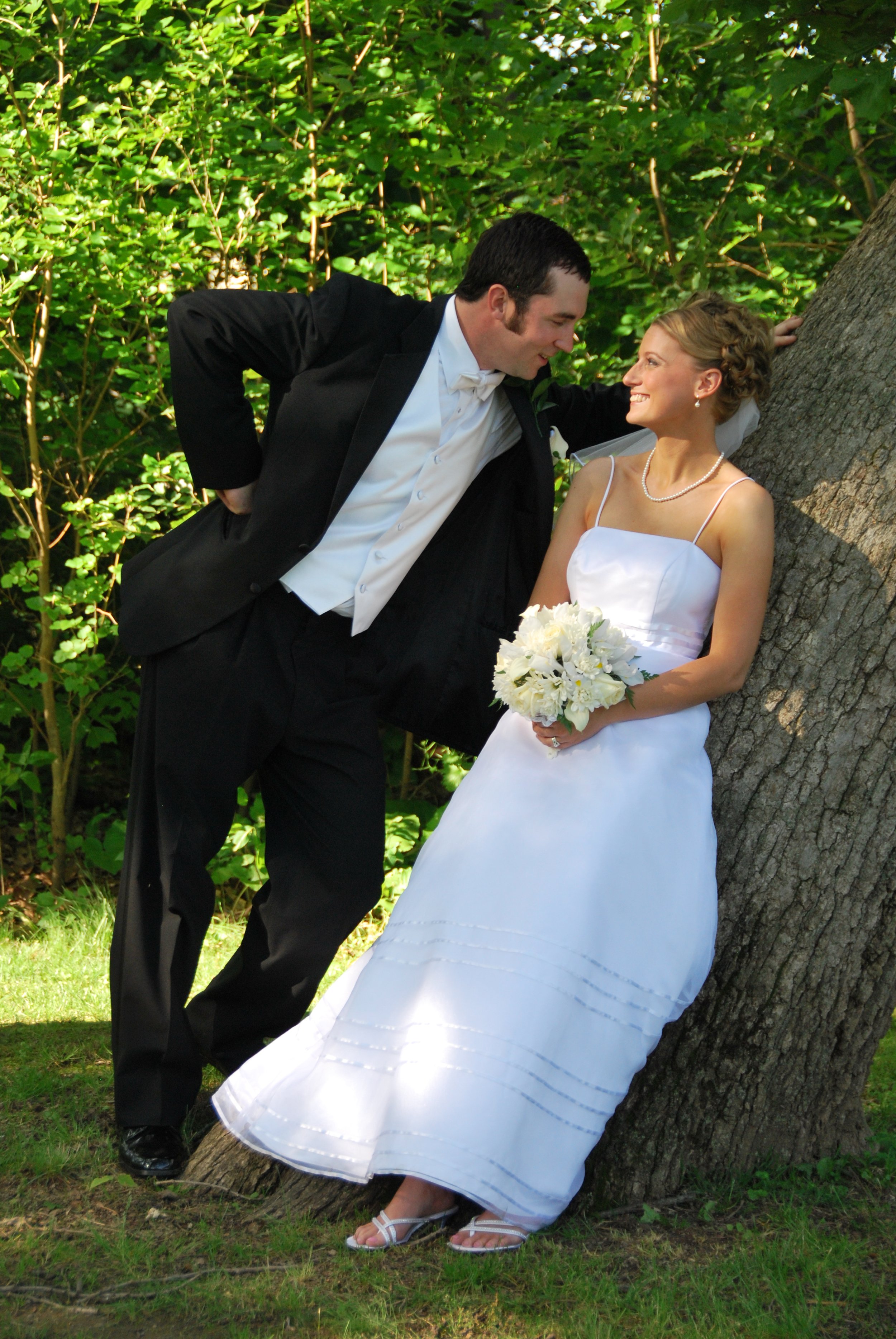 Bride and groom smiling at each other outdoors, the bride in a white dress holding a bouquet and the groom in a tuxedo leaning against a tree.