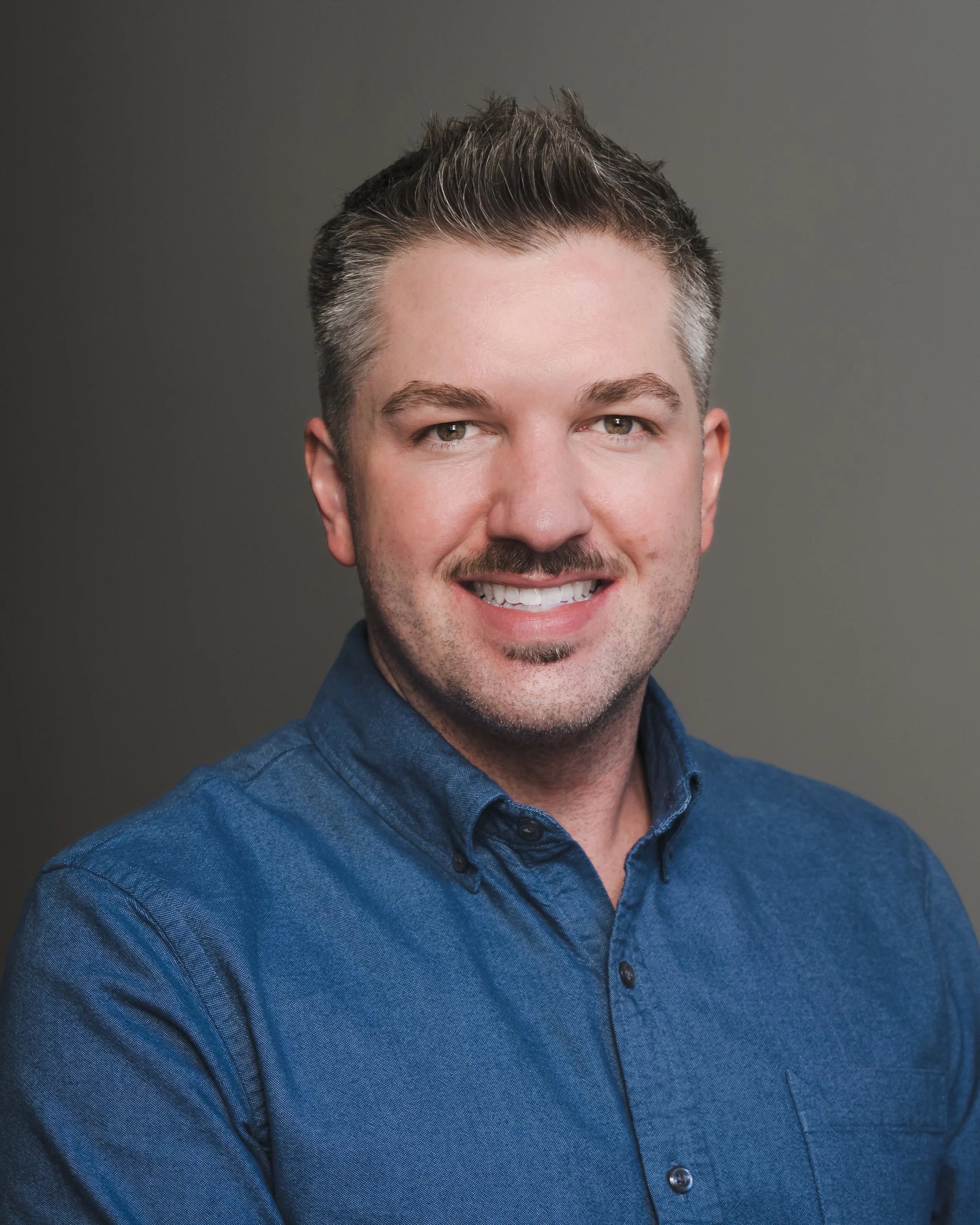 A man in a blue button-up shirt with short hair and a mustache, smiling against a neutral background.
