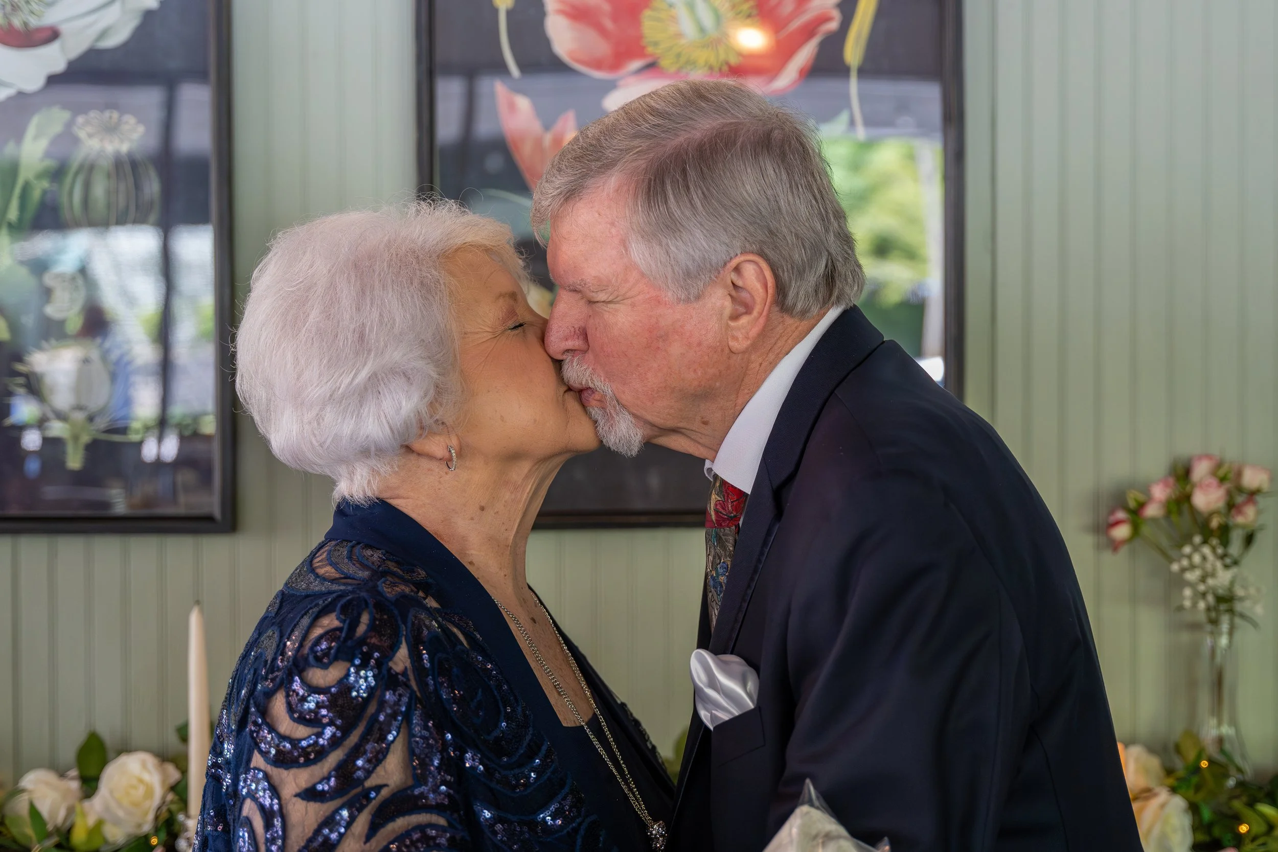 An elderly couple kissing affectionately in a room with floral decorations and framed art in the background.