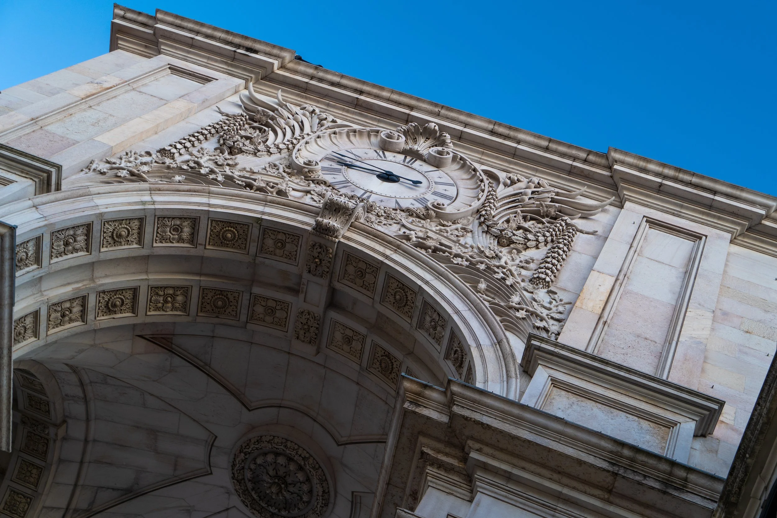 Architectural detail of an ornate stone arch with a decorative clock and sculptural elements, set against a clear blue sky.