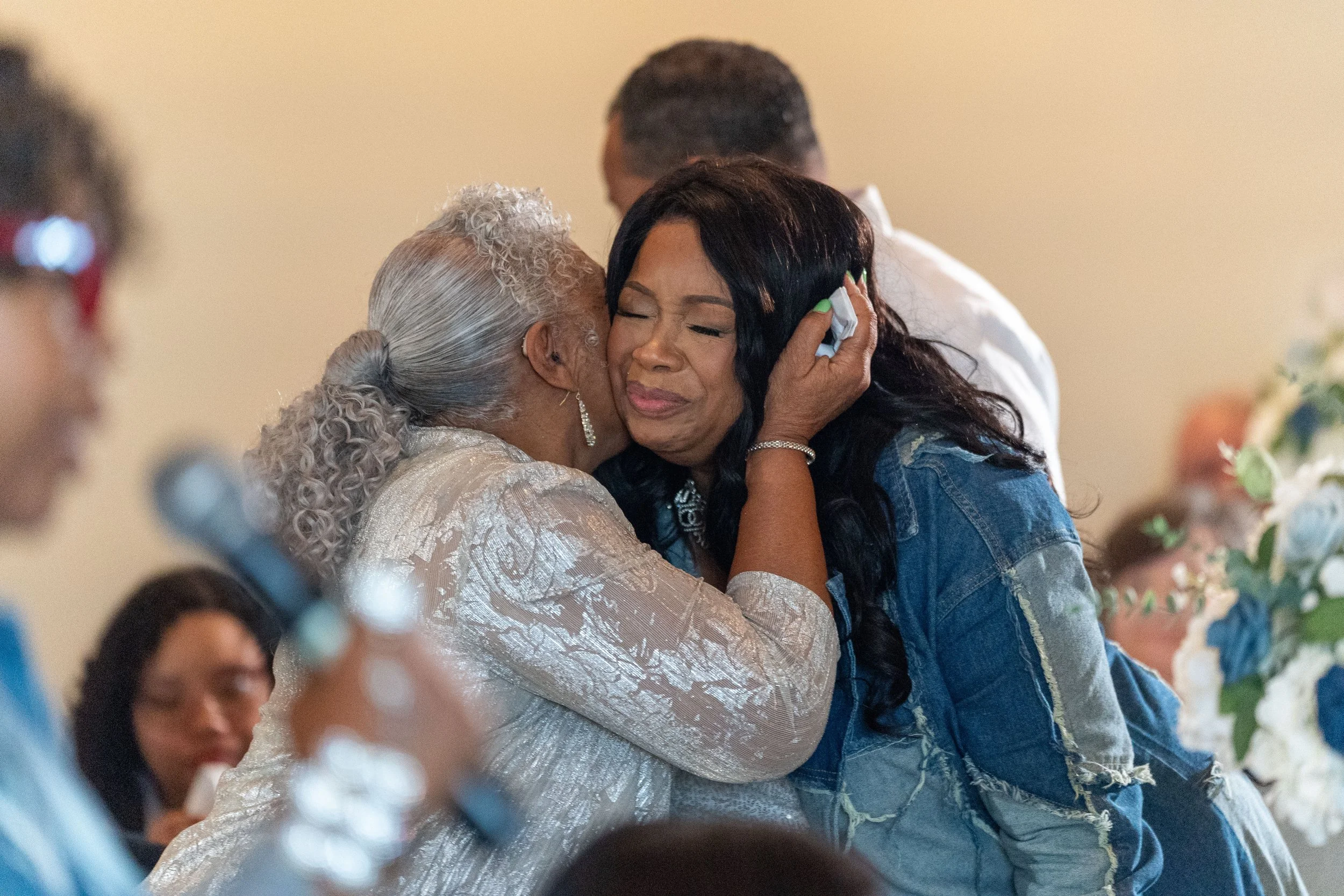 Two women hugging emotionally at a gathering, with a third person holding a microphone nearby.