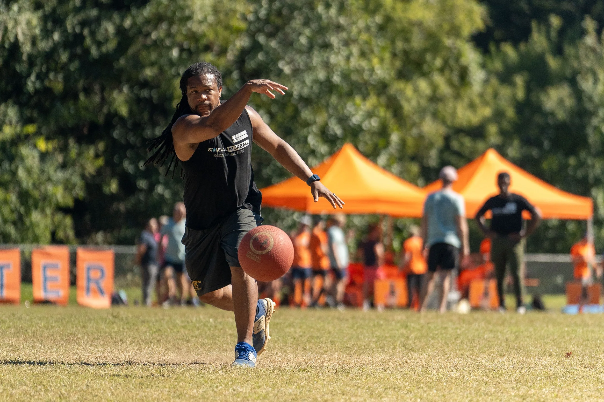 Person playing kickball on a grassy field, wearing a black tank top and shorts, surrounded by orange tents and spectators.