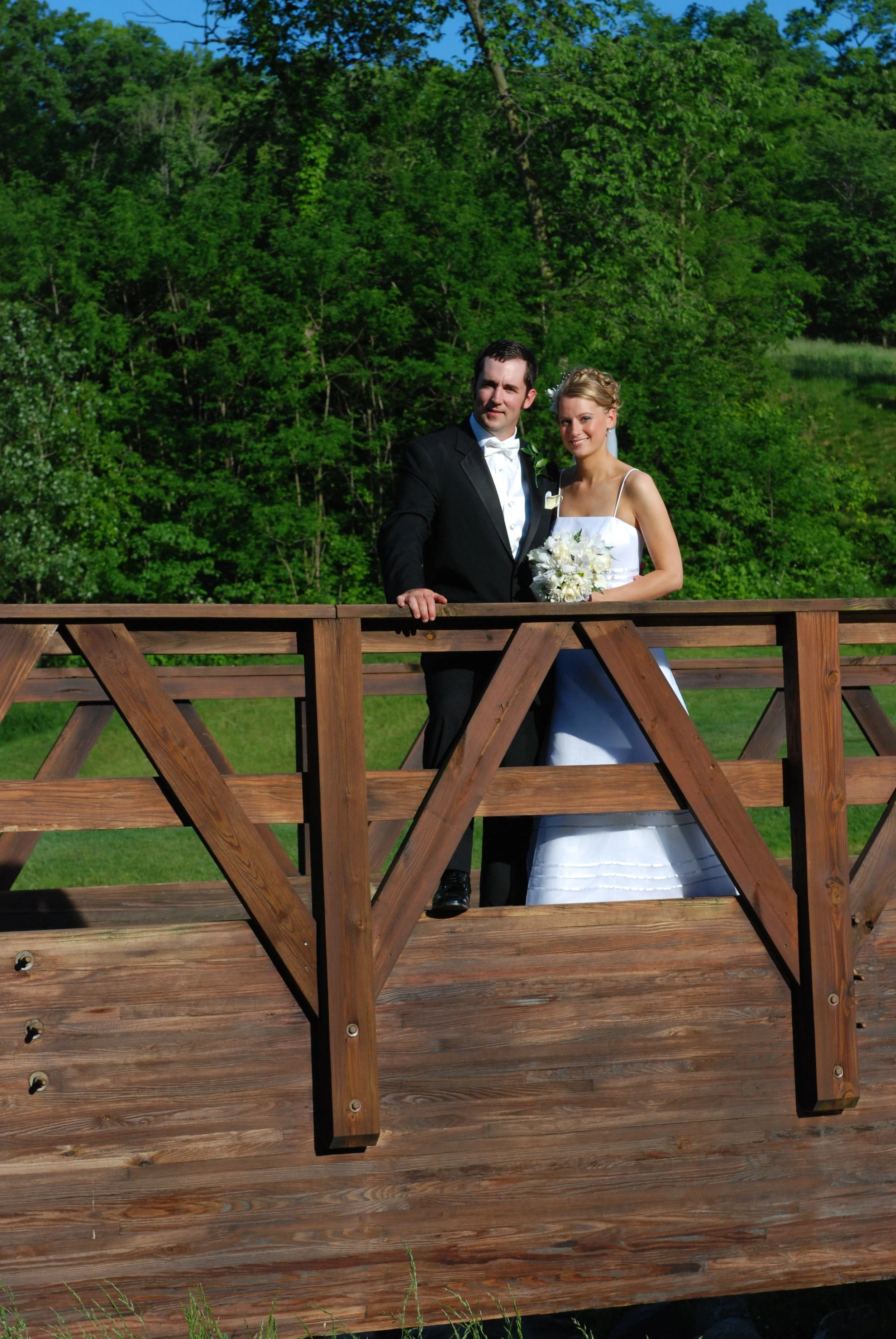 Bride and groom standing on a wooden bridge, surrounded by greenery, with the bride holding a bouquet.
