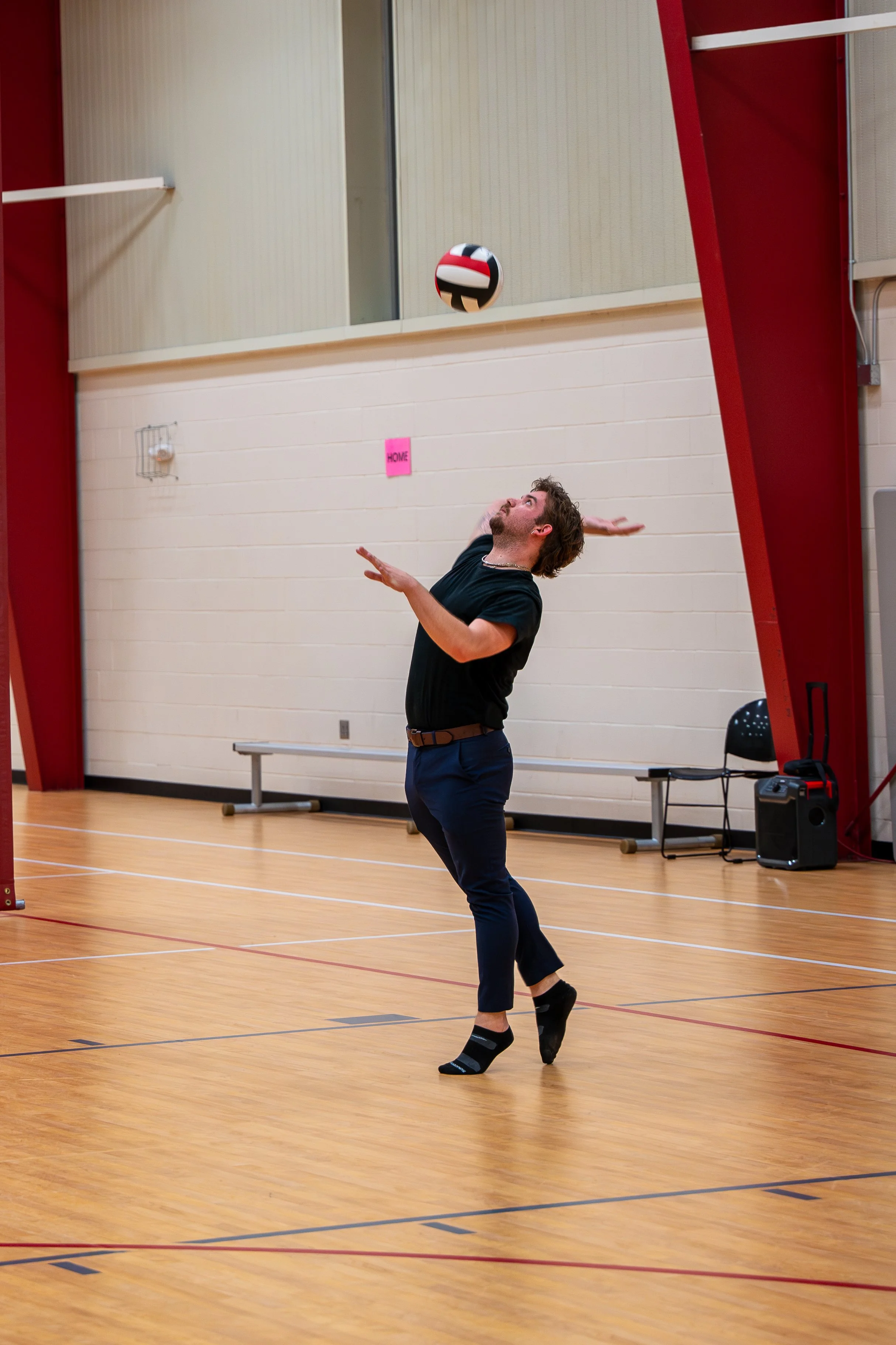 Person in a gymnasium performing a volleyball serve, wearing a black shirt and blue pants.