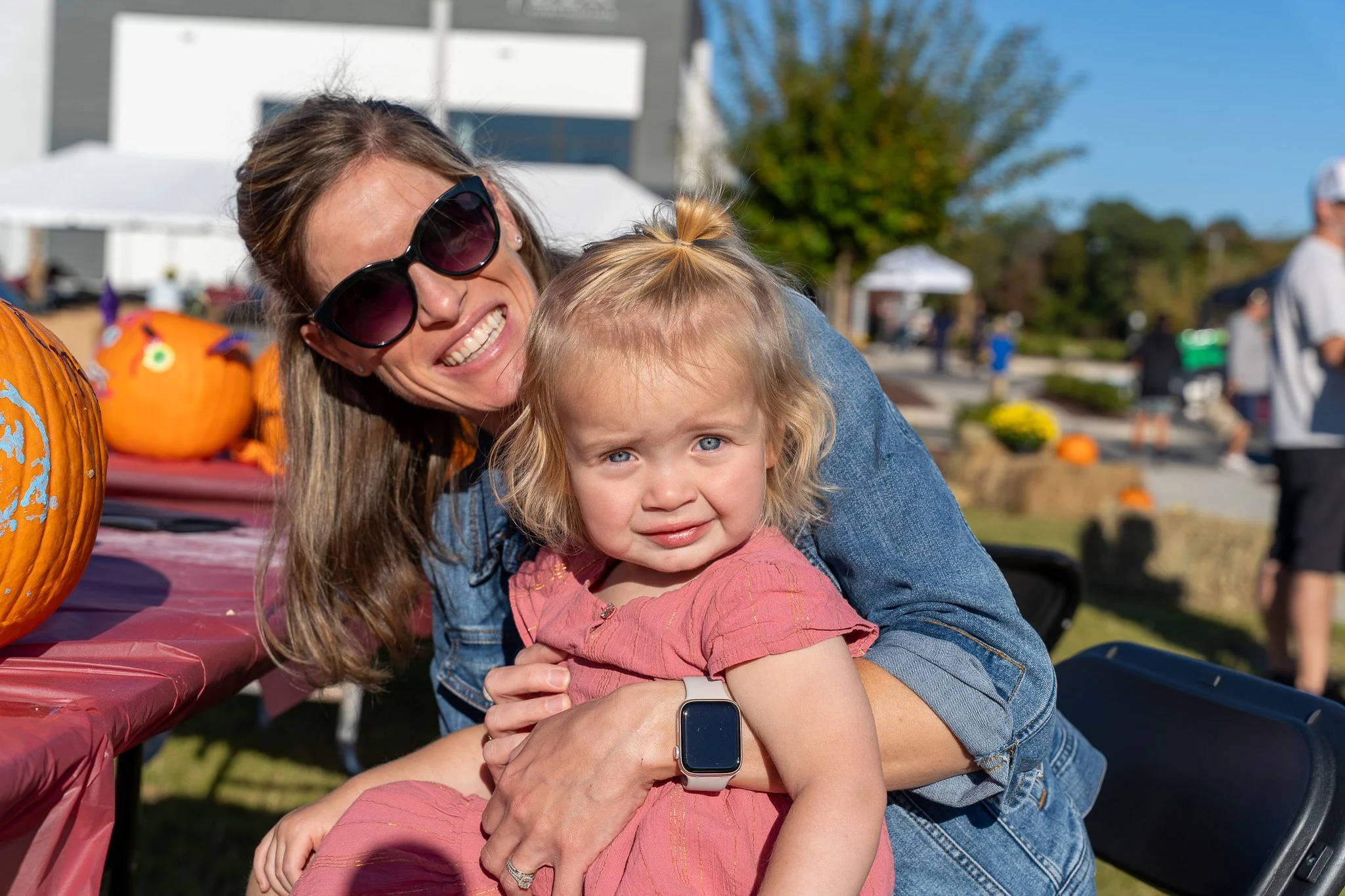 Smiling mother in sunglasses holding her young daughter at an outdoor pumpkin festival.