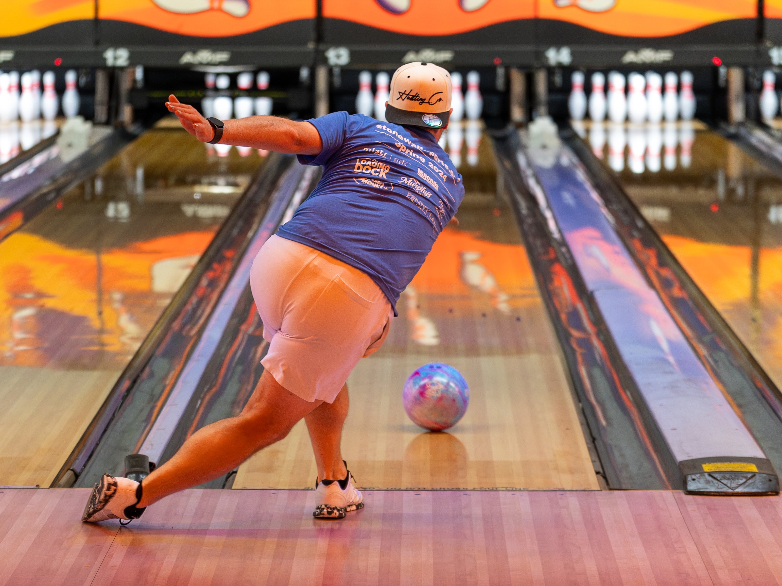 Bowler in motion rolling a bowling ball down the lane, wearing a blue shirt and white shorts, in a bowling alley with orange decor.