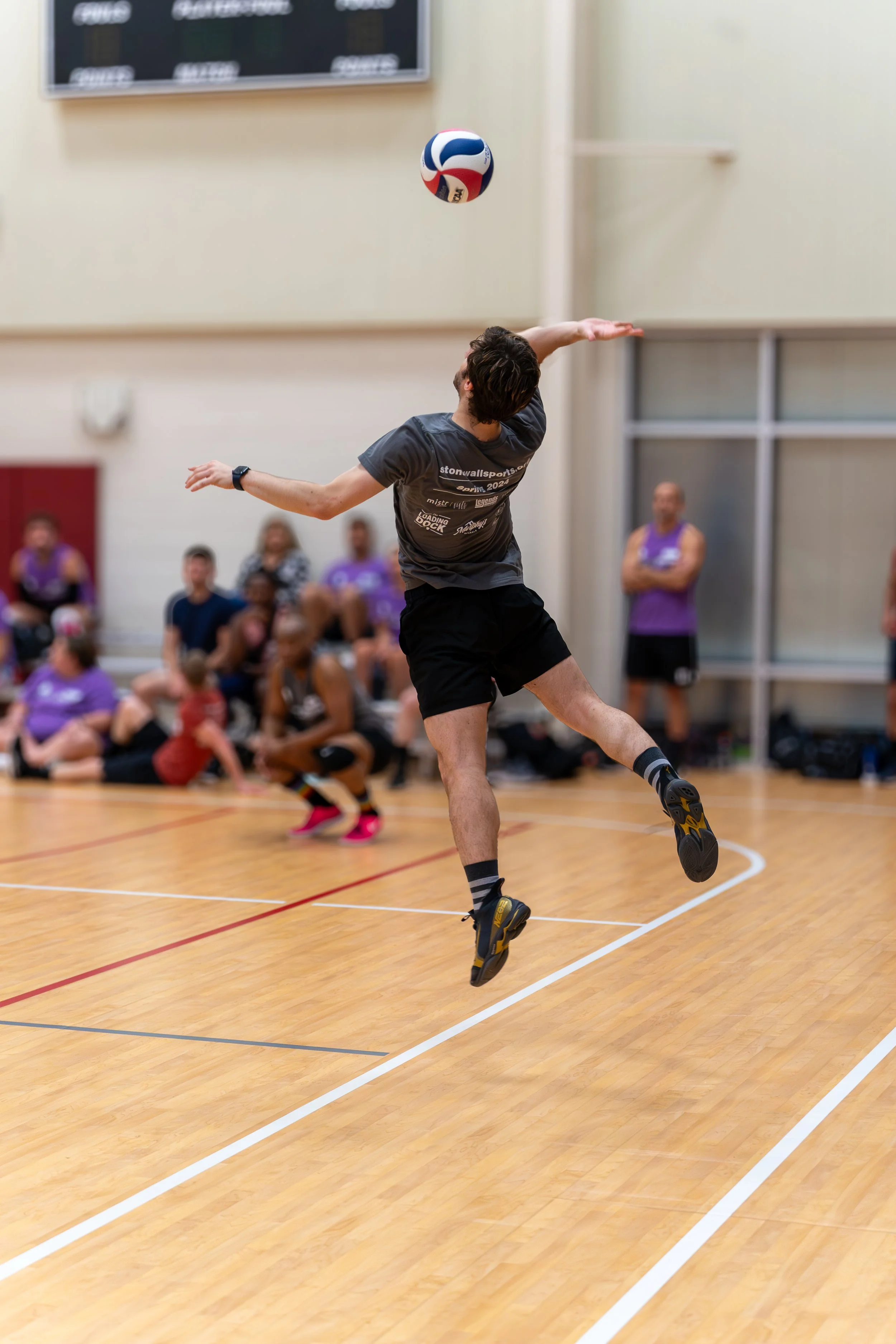A man performing a jump serve in an indoor volleyball game, with a volleyball in mid-air and spectators seated in the background.